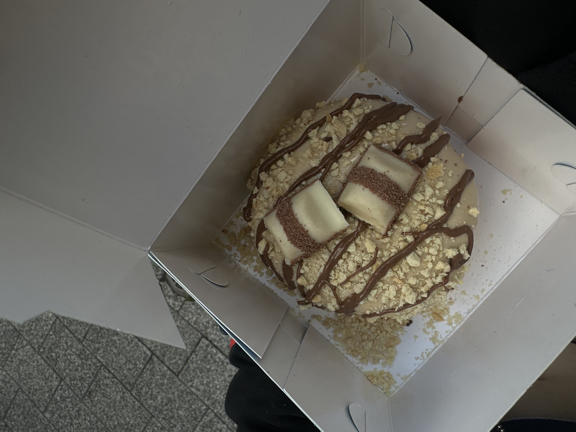 A donut is situated in an open white box. The donut has a light-colored glaze, crushed nuts, and chocolate drizzle, with two Kinder Bueno candy pieces placed on top. The box is sitting on what appears to be a stone-paved surface. A person's black-clad arm or leg is visible outside the box. The photograph appears to be taken in Plovdiv, Bulgaria.