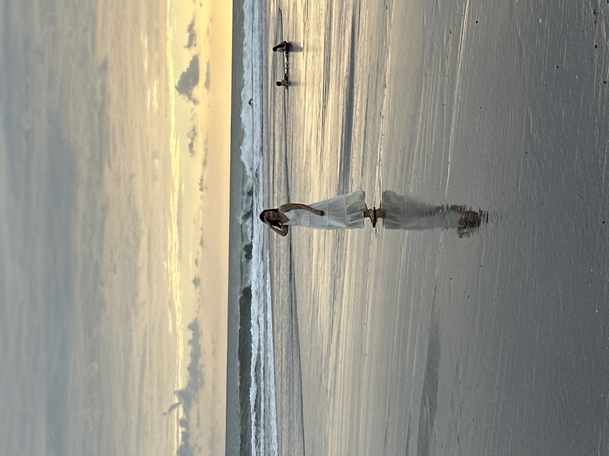In North Kuta, Indonesia, a young woman in a white sundress stands on a wet sand beach, her reflection shimmering on the ground. Her left hand is raised to her forehead, as if shielding her eyes from the sun, while her right hand rests on her hip. Behind her, the ocean churns with waves, and in the distance, two individuals are partially submerged in the water, their bodies suggesting they are either swimming or playing. The sky above is overcast with soft, muted clouds, with a pale yellow glow on the horizon, indicating either dawn or dusk. The overall scene depicts a serene beach environment.