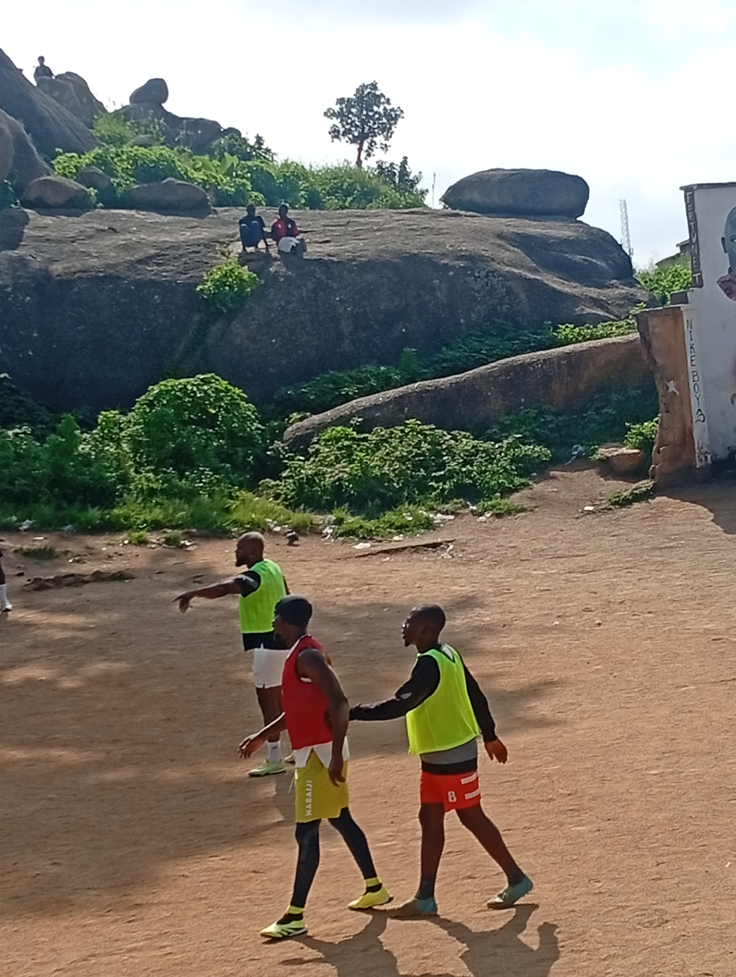 The image shows three young men in athletic wear on a dusty field, with large rock formations in the background. Two of the men are in the foreground, one wearing a red vest and yellow shorts, the other in a lime green vest and red shorts. The third man, in a lime green vest and white shorts, stands slightly behind and to the left. The rock formations appear to be a prominent natural feature, covered in green vegetation. In the far background, a sign with text that includes "NIKE BOYS" is visible on a wall. The setting suggests an outdoor sports or training area, possibly a park or a recreational space within Jos, Nigeria, known for its rocky terrain. The lighting indicates it is daytime, with clear skies.