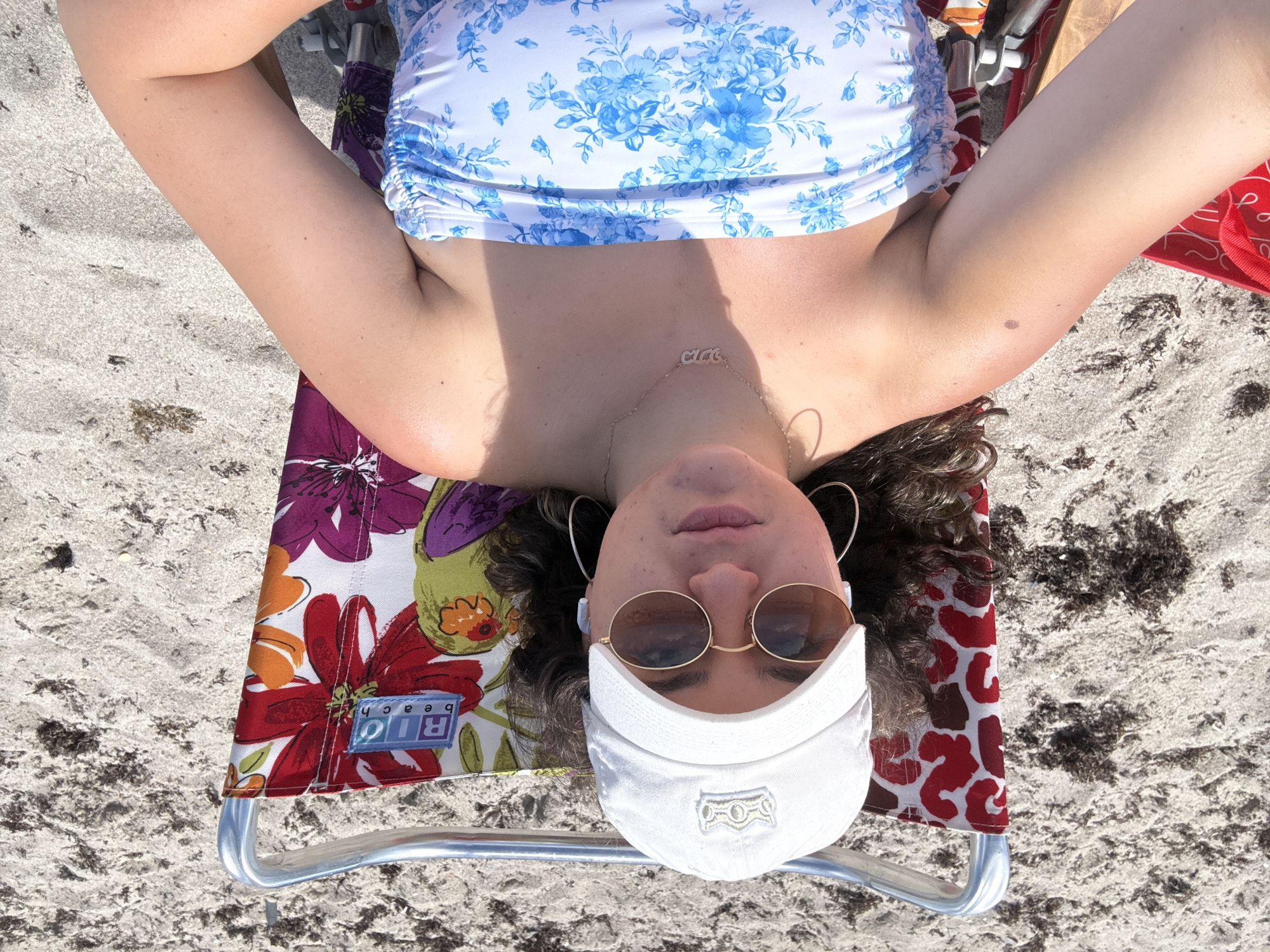 A woman is lying on a beach chair in Hollywood, Florida. The woman is wearing a blue floral top, sunglasses, and a white baseball cap. The beach chair has a colorful floral pattern on the seat and a red patterned back. The beach is sandy with some seaweed present. The photo is taken from an overhead perspective.