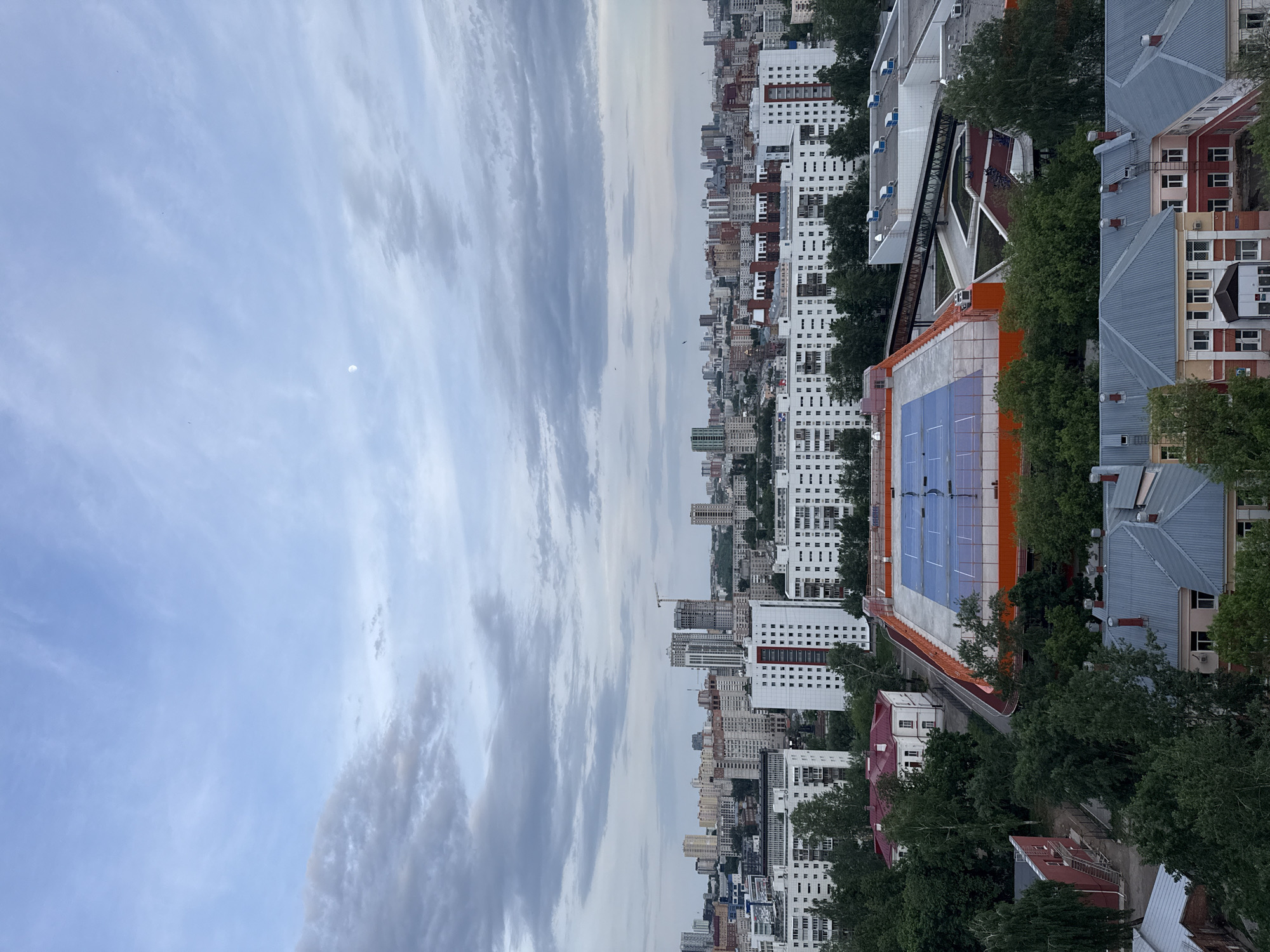 The image depicts a cityscape in Perm, Russia. High-rise apartment buildings dominate the skyline, primarily white with red and beige accents. A sports facility with orange walls and a blue rooftop is visible in the foreground, surrounded by trees. The sky is overcast, with cloud streaks running vertically. A small pale circle, potentially the moon, is visible in the sky. Other lower buildings with sloped roofs are adjacent to the high rises.