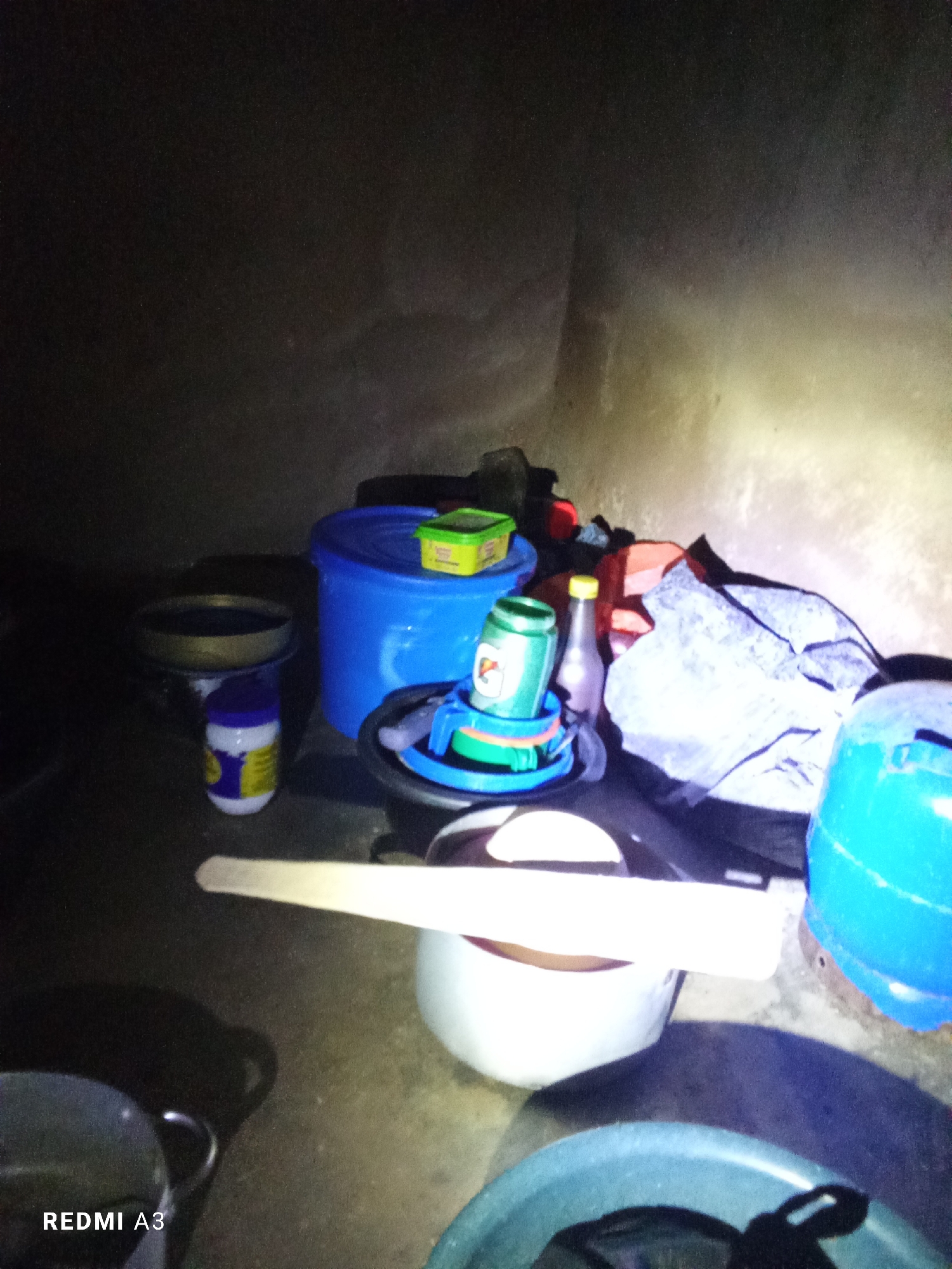 This is a dimly lit, indoor scene with a collection of household objects. In the center, a large blue bucket with a green lid sits on the floor. To its left, a smaller blue container with a yellow lid is visible, as is a white and purple bottle. A wooden utensil rests horizontally across a white pot in the foreground. To the right of the blue bucket, a large, crumpled gray bag is present, along with a bright blue container. A bottle with a yellow cap is also visible, situated between the blue bucket and the gray bag. The lighting suggests it might be nighttime or a poorly lit interior space. The overall impression is one of a cluttered storage area or a makeshift workspace. The text "REDMI A3" is visible in the bottom left corner of the image, indicating it is likely a photograph taken with a Redmi A3 phone.