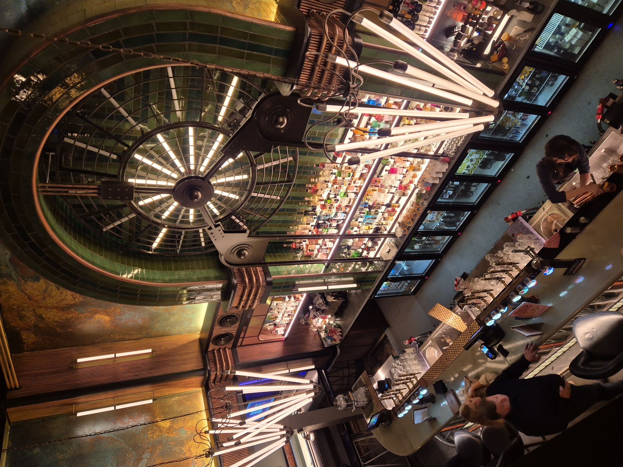 The image depicts an interior view of a bar, possibly in London, United Kingdom. The scene is oriented upward, showcasing the ceiling design, lighting, and the bar setup. A distinctive ceiling fixture with a radiating pattern and an outer ring structure dominates the upper left portion of the image. Rows of bottles are displayed on shelves, illuminated by linear light fixtures above. A section of the bar is visible along the right side, with a person standing behind the counter and another seated at the bar. Drinks are being prepared on the counter. There are multiple cold-display refrigerators filled with beverages. The bar counter has beer taps and other accessories. The environment is dimly lit, creating a modern ambiance.