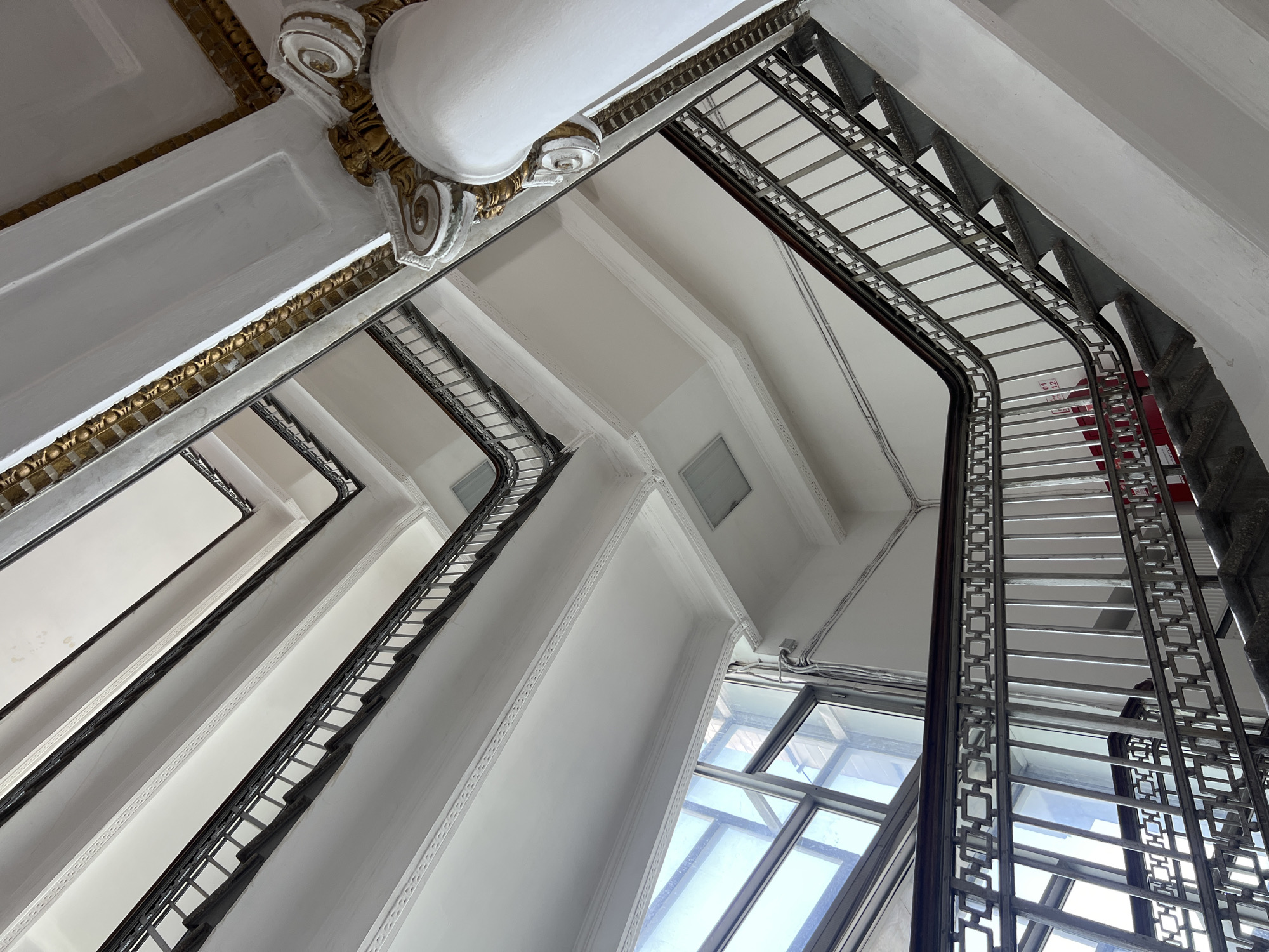 An upward-angled shot captures an ornate staircase within a building, possibly in Moscow, Russia. The stairs have metal railings with geometric designs. The walls are white with decorative trim, including gold accents in some areas. Large windows are visible, providing natural light. The architecture features strong vertical lines and repeating patterns.