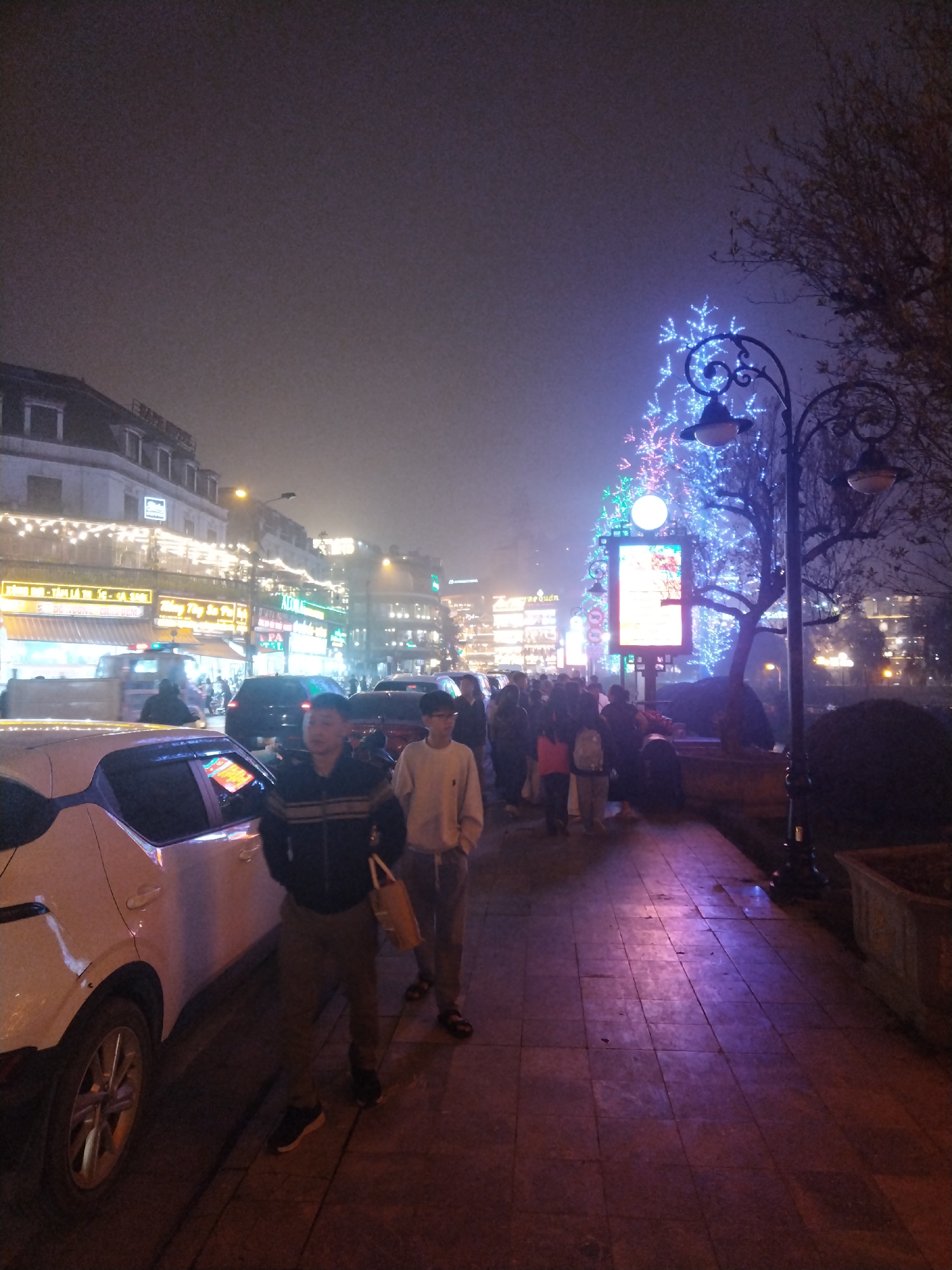 This nighttime image shows a street scene in Sa Pa Ward, Vietnam. The scene is illuminated by a mix of streetlights and colorful Christmas decorations. A Christmas tree decorated with lights is a central feature on the right side of the image. A crowd of people is walking along a brick-paved sidewalk. Two men are walking near a parked white car in the foreground. Buildings with shops and restaurants line the left side of the street, adorned with lights. The sky is dark and appears foggy.
