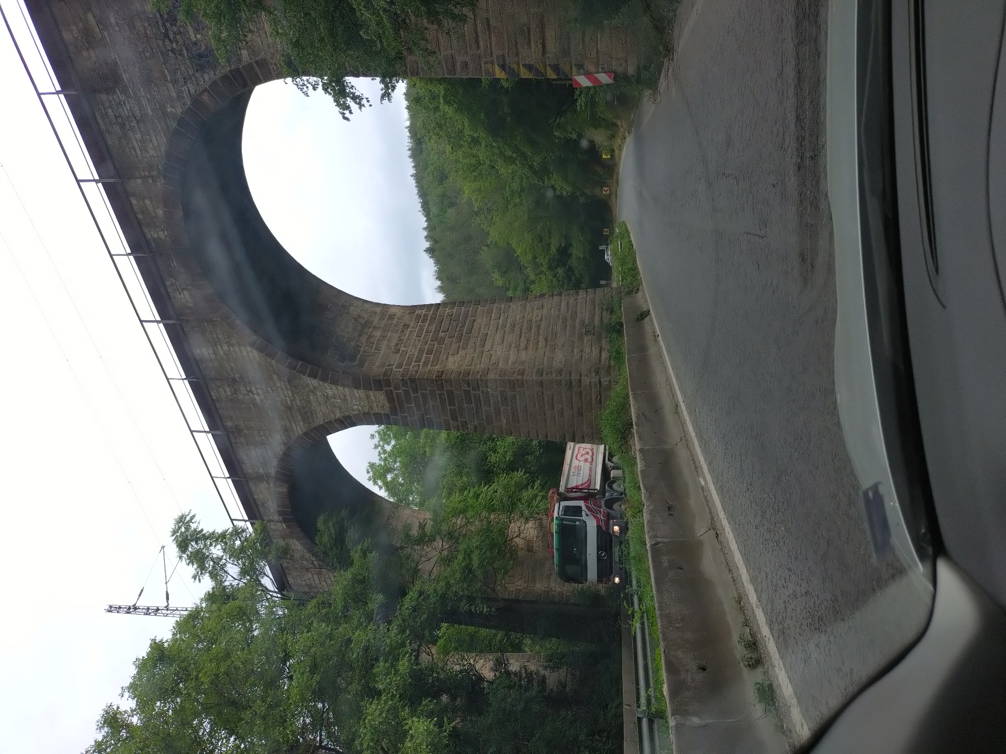 An eye-level, slightly wide-angle shot captures a view from a vehicle looking forward, towards a large stone arched viaduct. The viaduct, constructed from rough-hewn stone blocks, dominates the upper portion of the frame, with two prominent arches visible. A railway track with overhead catenary is present on top of the viaduct.
Below the viaduct, a two-lane asphalt road curves gently to the right and disappears into a wooded hillside. On the left side of the road, a white and red truck is driving, partially obscured by the viaduct's arch. The truck has "SS" logo on its side. A concrete barrier runs along the left edge of the road.
The environment is lush and green, with dense foliage and trees flanking the road and surrounding the viaduct. The sky is overcast and light gray. The scene appears to be a road passing under a railway viaduct, surrounded by nature. No people are directly visible in the image, except for the driver of the vehicle from which the photo is taken.
The location is the railway viaduct near the city of Zmeyovo, Bulgaria.