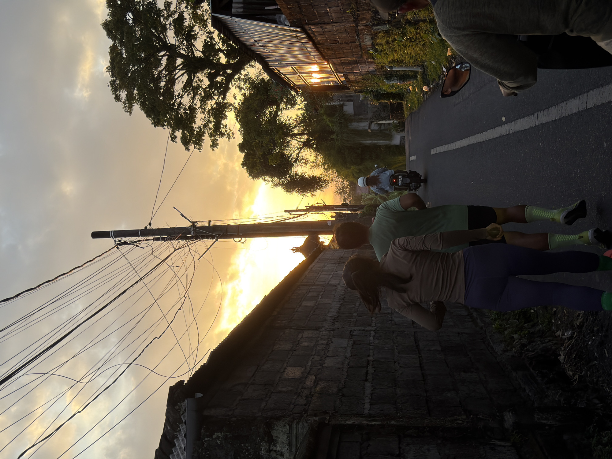 This image depicts an early morning or late afternoon scene on a street in North Kuta, Indonesia. The sky is filled with warm, golden hues of sunset or sunrise, with scattered clouds. A prominent utility pole with numerous tangled wires runs across the upper portion of the frame. On the left, a dark, textured brick wall lines the street. On the right, buildings with corrugated metal roofs and stone walls are visible, with lush green vegetation growing along their sides. The street itself is paved with asphalt and marked with a white line.
In the foreground, two individuals are seen running away from the camera. The person on the left has dark hair tied in a ponytail and is wearing a light brown long-sleeved shirt and dark purple leggings, with bright green socks and dark shoes. The person on the right is wearing a green t-shirt and black shorts, with light green socks and dark shoes.
Further down the street, a person is riding a black motorcycle with a white helmet. Another person is seated on a scooter or motorcycle in the foreground, seen from the back and partially reflected in the side mirror, wearing a beige top. The overall atmosphere suggests a casual, active moment within a residential or semi-urban setting in Indonesia.