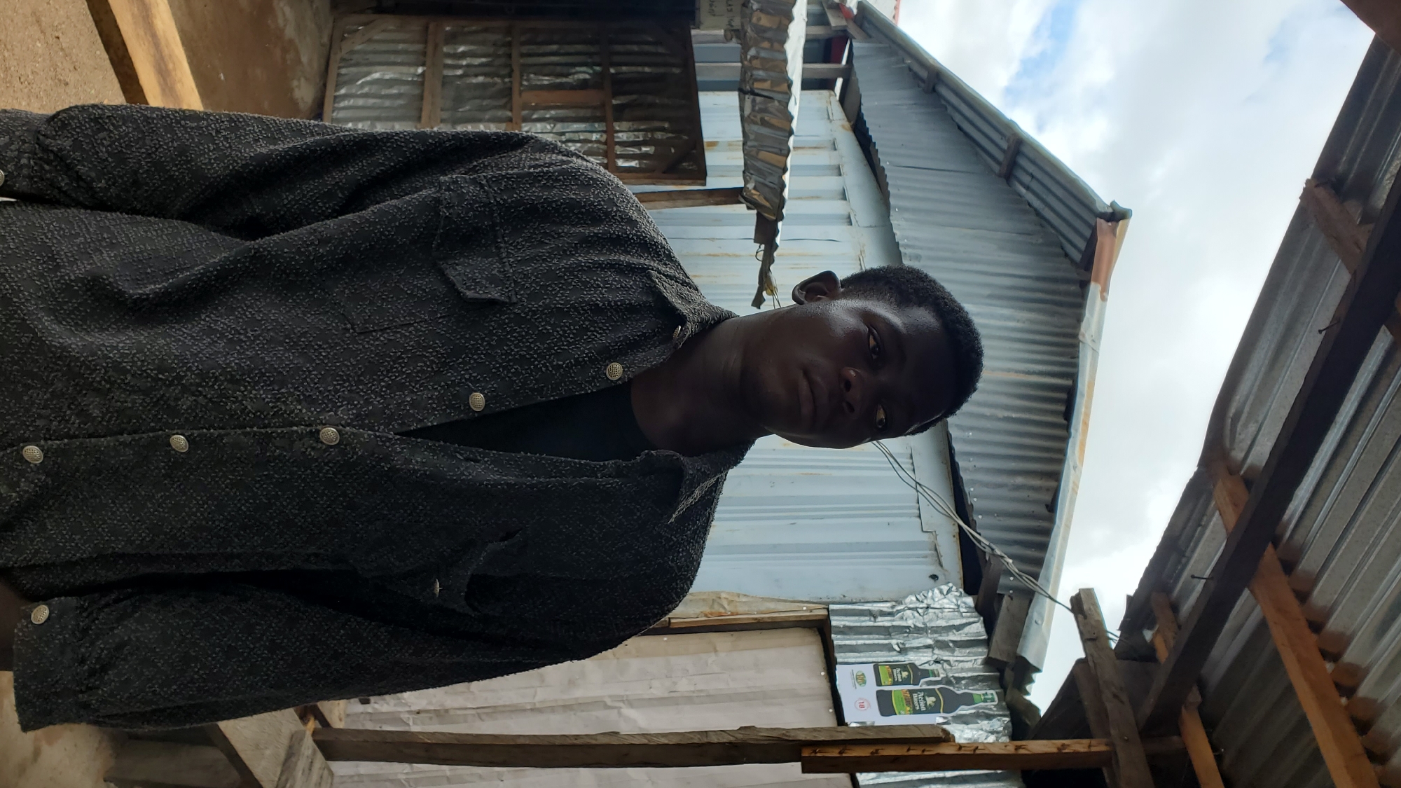 A young Black male stands in front of a corrugated metal structure, possibly a shop or dwelling, in Jalingo, Nigeria. He is wearing a dark, textured collared shirt over a black t-shirt. The background consists of various corrugated metal walls and roofing, with wooden supports visible. A poster featuring two bottles of alcohol is affixed to the right side of the structure. The sky above is overcast.