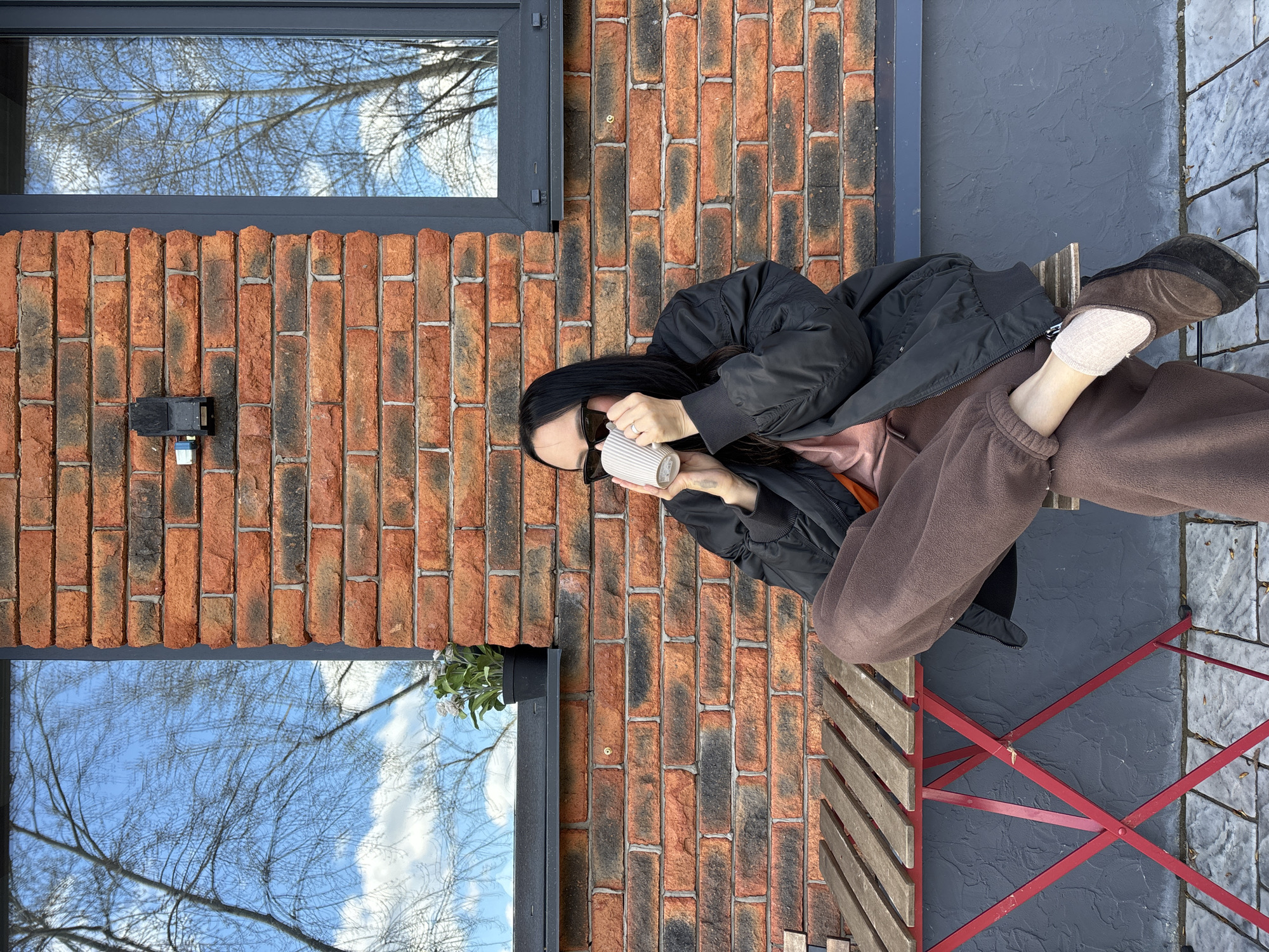 A woman wearing sunglasses, a black jacket, brown sweatpants, and slippers sits on a wooden chair outside a brick building. She holds a white mug in both hands. The building has two windows reflecting a blue sky with bare tree branches. A black surveillance camera is mounted on the brick wall between the windows. The chair has a red metal frame and is placed on a textured gray surface resembling paving stones. The scene is located in the city of Chaikovsky, Russia.