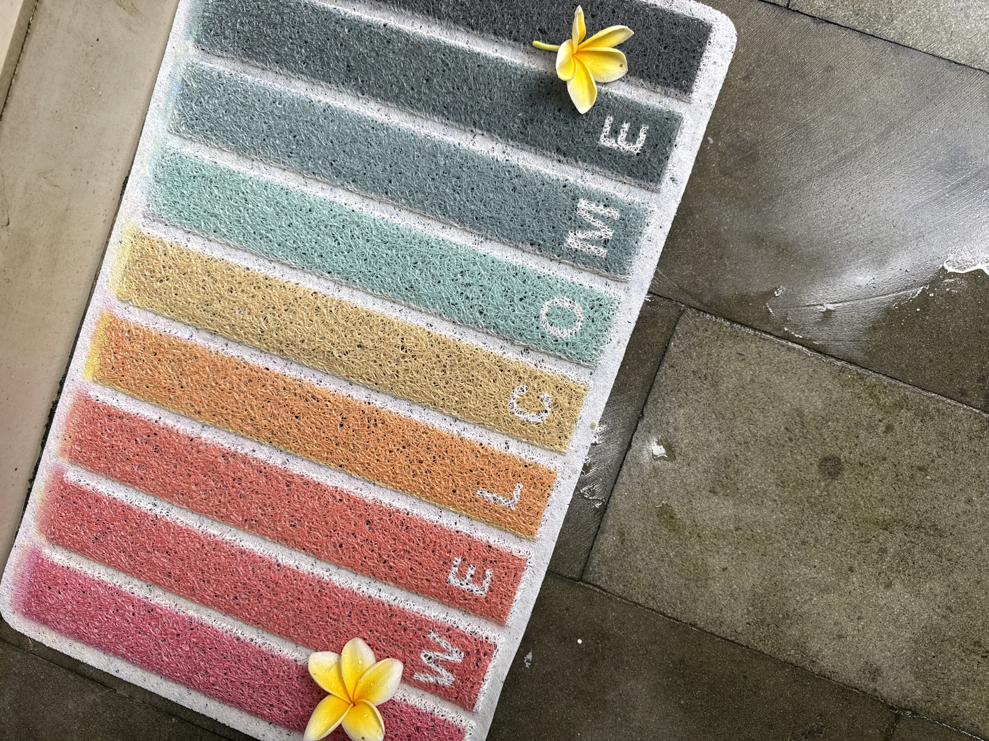 An overhead, slightly angled shot captures a vibrant "WELCOME" doormat laid on a textured concrete or tiled surface. The doormat features seven horizontal stripes of looped PVC strands, each in a distinct color. From top to bottom, the stripes are dark gray, teal, light blue, beige, orange, reddish-orange, and pink. The word "WELCOME" is spelled out vertically in prominent white capital letters, with each letter centered on its respective stripe.
Two yellow and white plumeria (frangipani) flowers are placed on the doormat: one rests on the top dark gray stripe, near the letter 'E', and the other is on the bottom pink stripe, beside the letter 'W'. The ground surrounding the doormat is composed of large, rectangular, dark gray concrete or stone tiles. Several sections of the ground, particularly to the right of the doormat, are visibly wet and reflective, with water droplets, indicating recent rain or a hosing down. The doormat itself also appears slightly damp.
The scene suggests an outdoor or entryway setting in a tropical environment, consistent with the Kuta Selatan, Indonesia context, reinforced by the presence of plumeria flowers. There are no people visible, and no specific activity is taking place; it's a static composition highlighting the doormat and its surroundings.