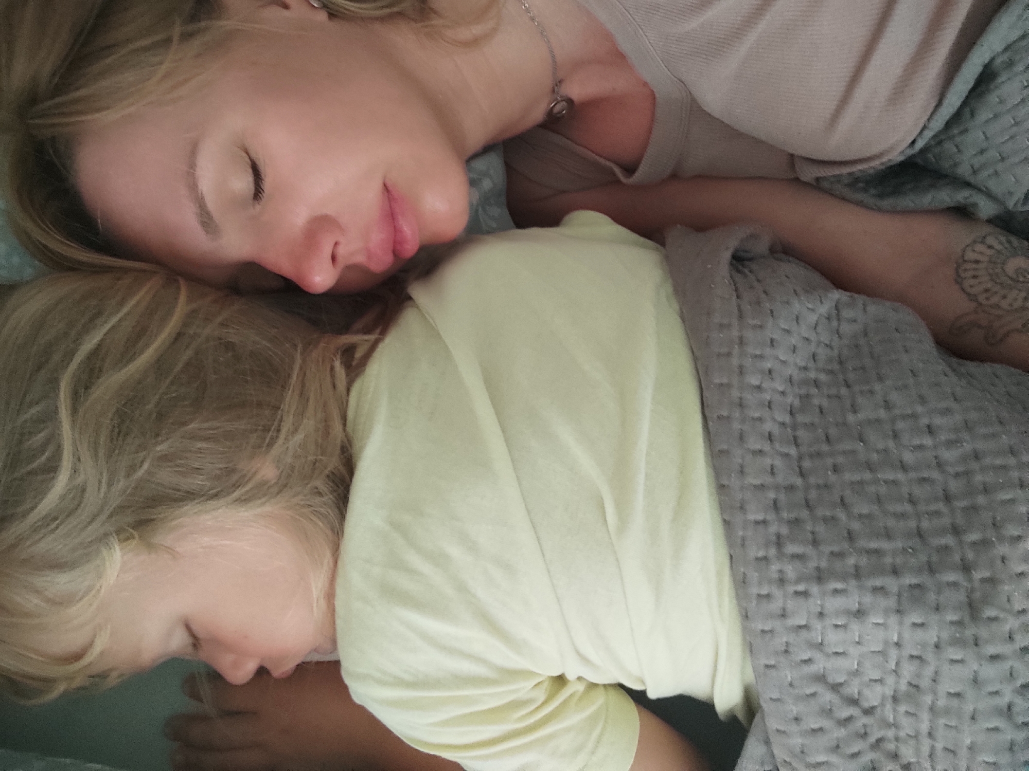 The image shows a close-up, top-down view of two blonde individuals, presumed to be a mother and child, sleeping together in a bed. The adult on top is lying on their left side, facing towards the left of the frame, with their eyes closed and head resting on a pillow. They are wearing a light-colored top with a silver necklace. The child is lying below the adult, also on their left side, with their head turned downwards and eyes closed. They are wearing a pale yellow t-shirt. Both individuals are covered by a grey quilted blanket. The environment appears to be a bedroom, with soft lighting. The specific location in Blanes, Spain is not discernible from the image itself.