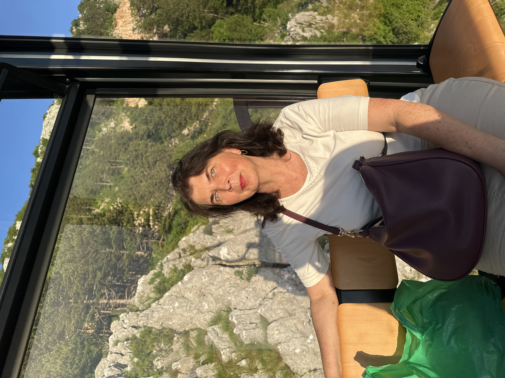A woman is seated in a cable car with a scenic mountain view. She has medium-length dark hair, blue eyes, and is wearing a white t-shirt and light-colored pants. She is carrying a dark purple handbag and a green plastic bag is next to her. The cable car's windows offer a view of rocky terrain and lush green trees under a clear blue sky. The lighting suggests it is late afternoon or early evening. The location is the cable car in Dub, Montenegro, which connects the town of Kotor to the village of Njeguši, offering panoramic views of the Bay of Kotor.