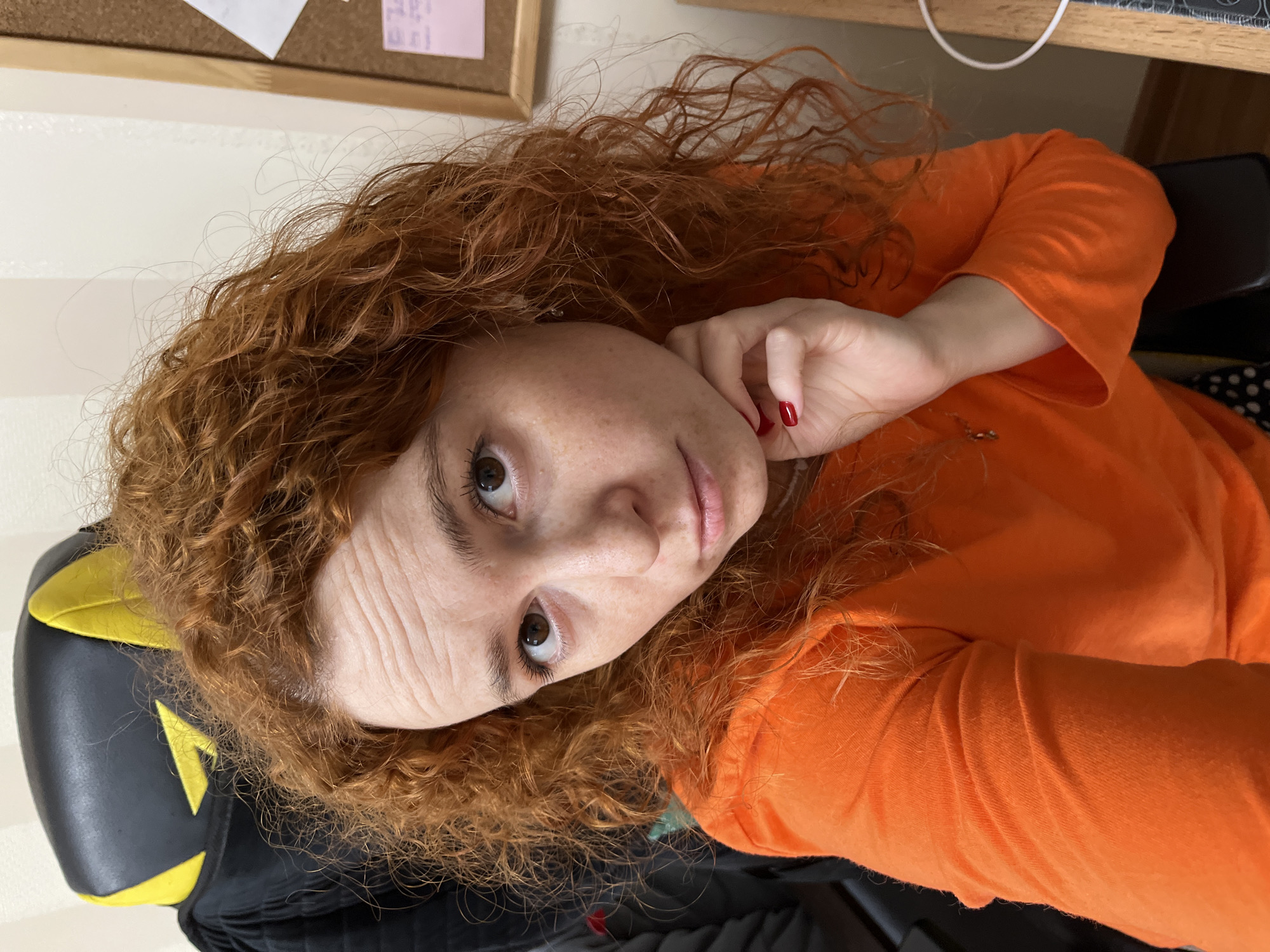 The image shows a woman with curly red hair and freckles, wearing an orange long-sleeved shirt. She is sitting on a black and yellow chair. Her right hand is near her face. A bulletin board is visible in the background. The exact location in Chaikovsky, Russia cannot be determined from the provided image alone.