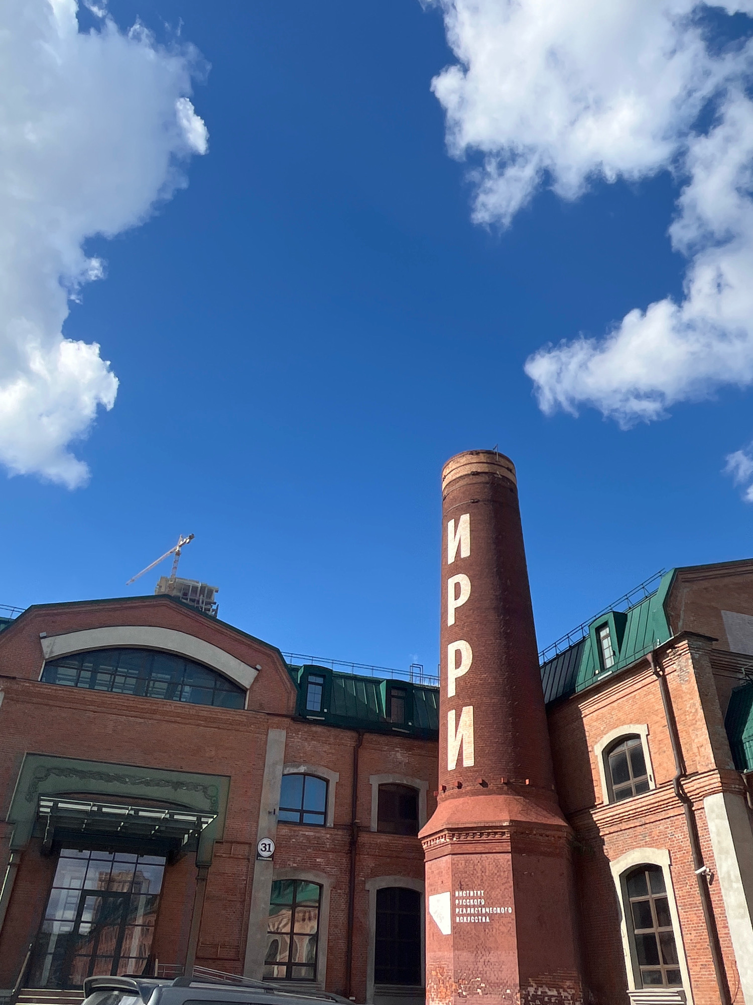 The image depicts the historic Ivanovo-Serebryakov Textile Factory building in Moscow, Russia, under a bright blue sky with scattered white clouds. A tall, reddish-brown brick chimney dominates the right side of the frame, bearing white Cyrillic lettering that reads "ИРРИ" vertically, and "ИНСТИТУТ РУССКОГО РЕАЛИСТИЧЕСКОГО ИСКУССТВА" at its base.
The main building is constructed of red brick with a green metal roof. The architecture features large arched windows and smaller rectangular windows. A prominent entrance with a glass canopy is visible on the left side, with a sign indicating "31" next to a window on the right side of the building. A construction crane can be seen in the distance behind the building, suggesting ongoing development or renovation. The foreground shows the roofline of a parked vehicle. The scene is a clear, sunny day, highlighting the industrial heritage and artistic presence of the location.