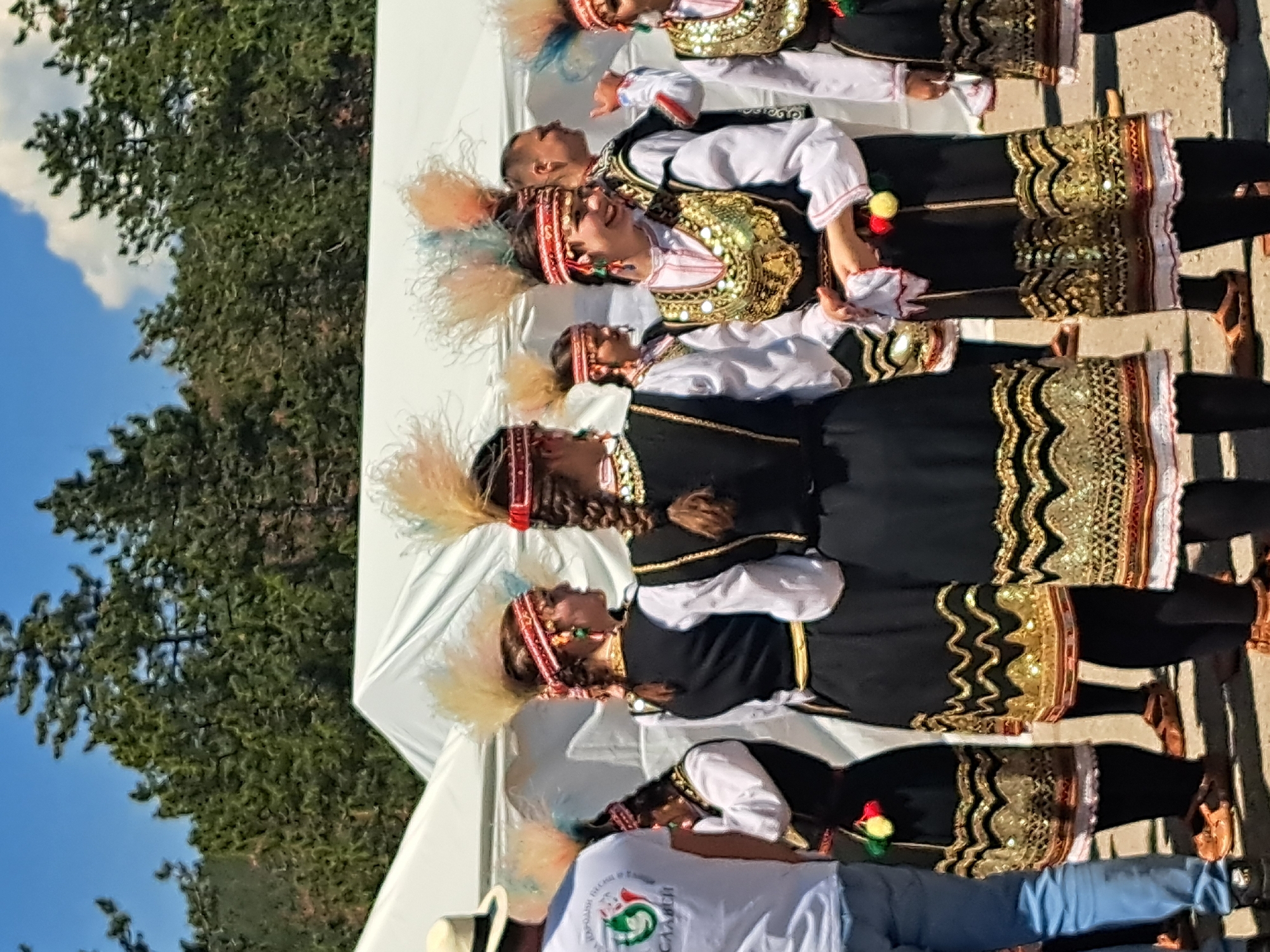 A group of people in traditional Bulgarian folk costumes stand in a line. They are wearing feathered headpieces, white shirts, and black vests and skirts with gold embroidery. The women are wearing leggings. The person in the front is wearing a white t-shirt with an emblem and blue jeans. A white tent is in the background. Behind the tent are trees and a blue sky with clouds. The location appears to be outdoors in the city of Unknown, Unknown. They may be performing a traditional Bulgarian dance.
