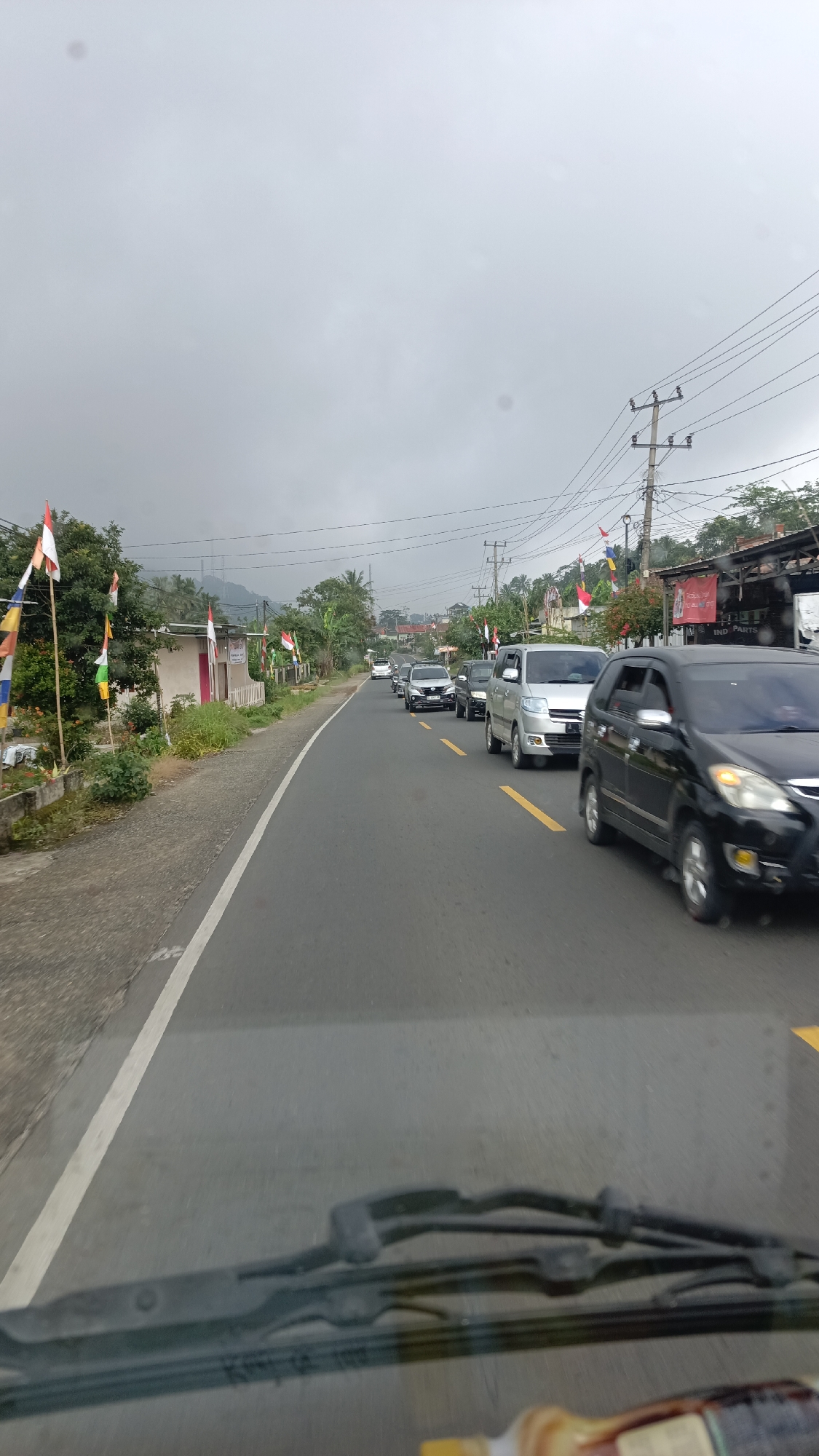 The image depicts a congested road in Sukabanjar, Indonesia, on an overcast day. A line of vehicles, including cars and vans, are visible on the asphalt road, moving slowly. The road is marked with yellow lines. On the left side of the road, there are houses, trees, and decorative flags, some of which appear to be national flags with red and white colors. Utility poles with power lines are present on the right side of the road, along with some buildings, one of which has a sign that says "IND PARTS". The foreground of the image shows the windshield and wipers of a vehicle, suggesting the photo was taken from inside a moving car. The overall scene indicates a typical traffic situation in a populated area, possibly during a festive period given the flags.