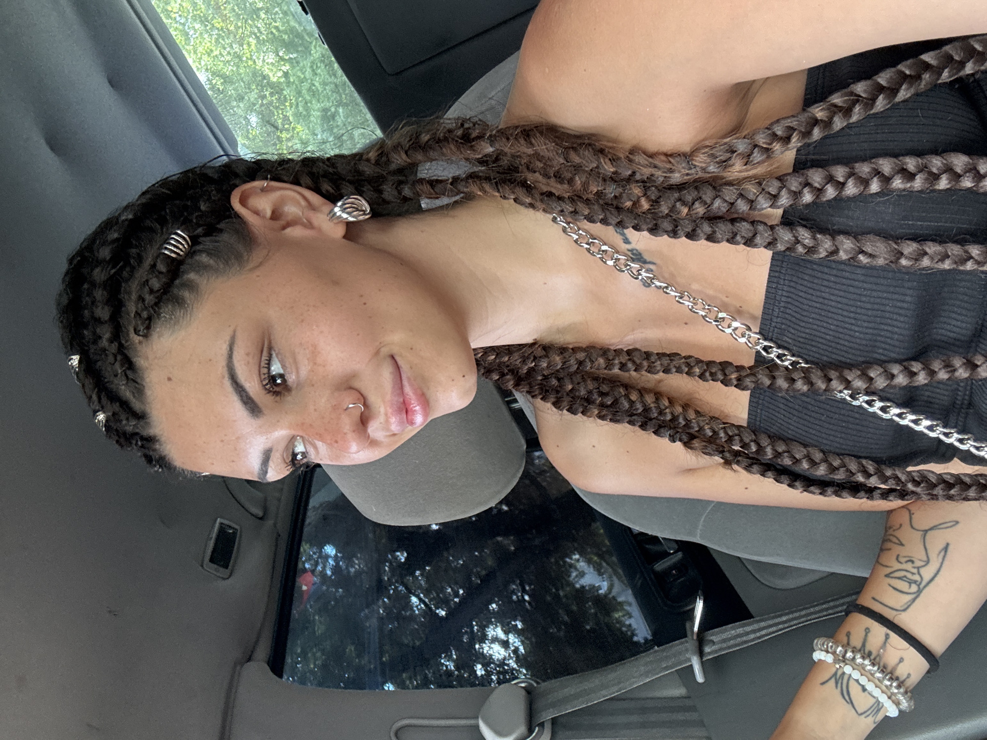 The media file is a portrait of a young woman with dark braided hair, wearing a black tank top and silver jewelry, including earrings, a nose ring, and a chain necklace. She has a tattoo of a face outline on her arm and is wearing bracelets. She is sitting in what appears to be the driver's seat of a car, indicated by the seatbelt and car interior. The background shows trees and foliage, suggesting an outdoor setting. The lighting indicates it is daytime. The woman is looking towards the camera with a slight smile. The overall mood is casual and relaxed. The location is identified as Sofia, Bulgaria.