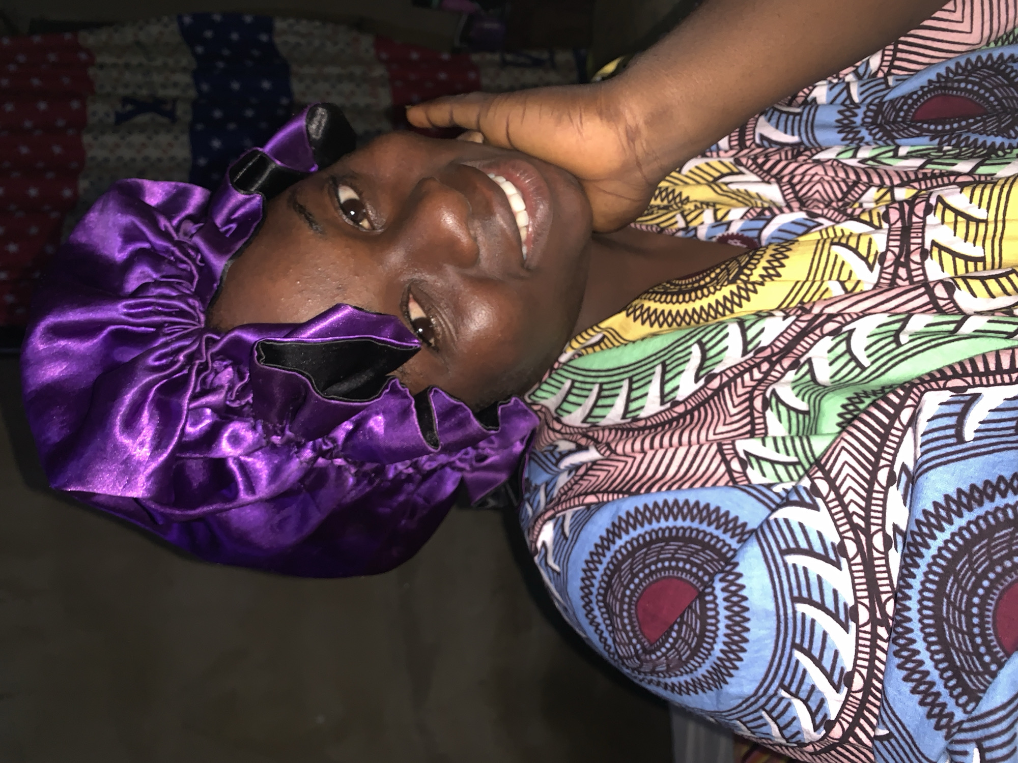 A dark-skinned woman wearing a purple satin bonnet and a colorful patterned shirt is shown in a close-up shot. Her right hand is resting on her chin, and she is smiling at the camera. The background is dark and nondescript. The image was taken in Agban, Nigeria.
