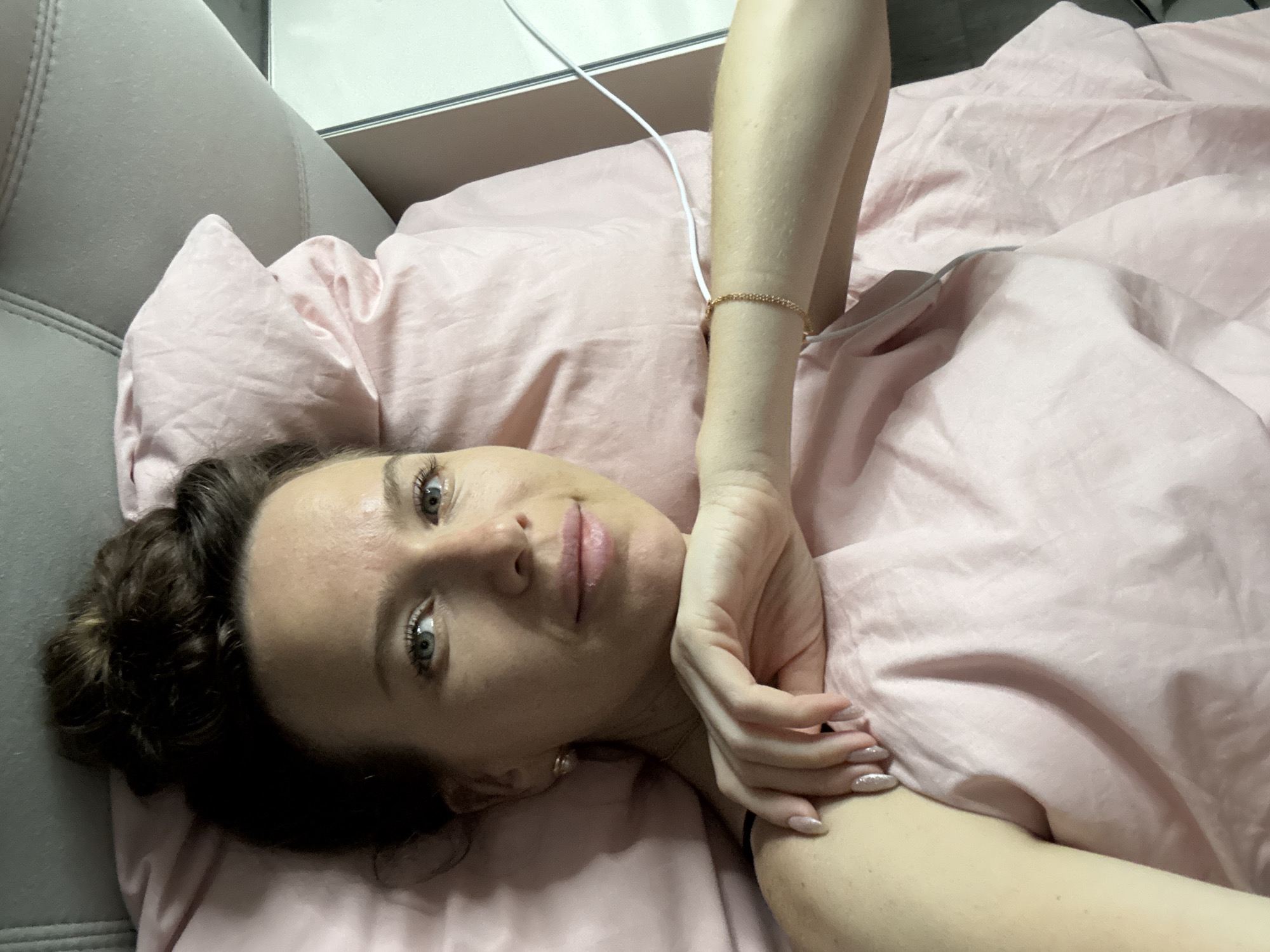 A woman with her hair in a bun, looking at the camera, lies on a pink duvet. She is wearing a black top and has her left arm bent, with her hand resting near her face. A gold bracelet adorns her wrist, and her fingernails are painted a shimmery pink. A thin white cord runs from behind her, across the duvet, and out of frame to the right. The background consists of a gray headboard and a pink pillow. The scene appears to be in a bedroom.