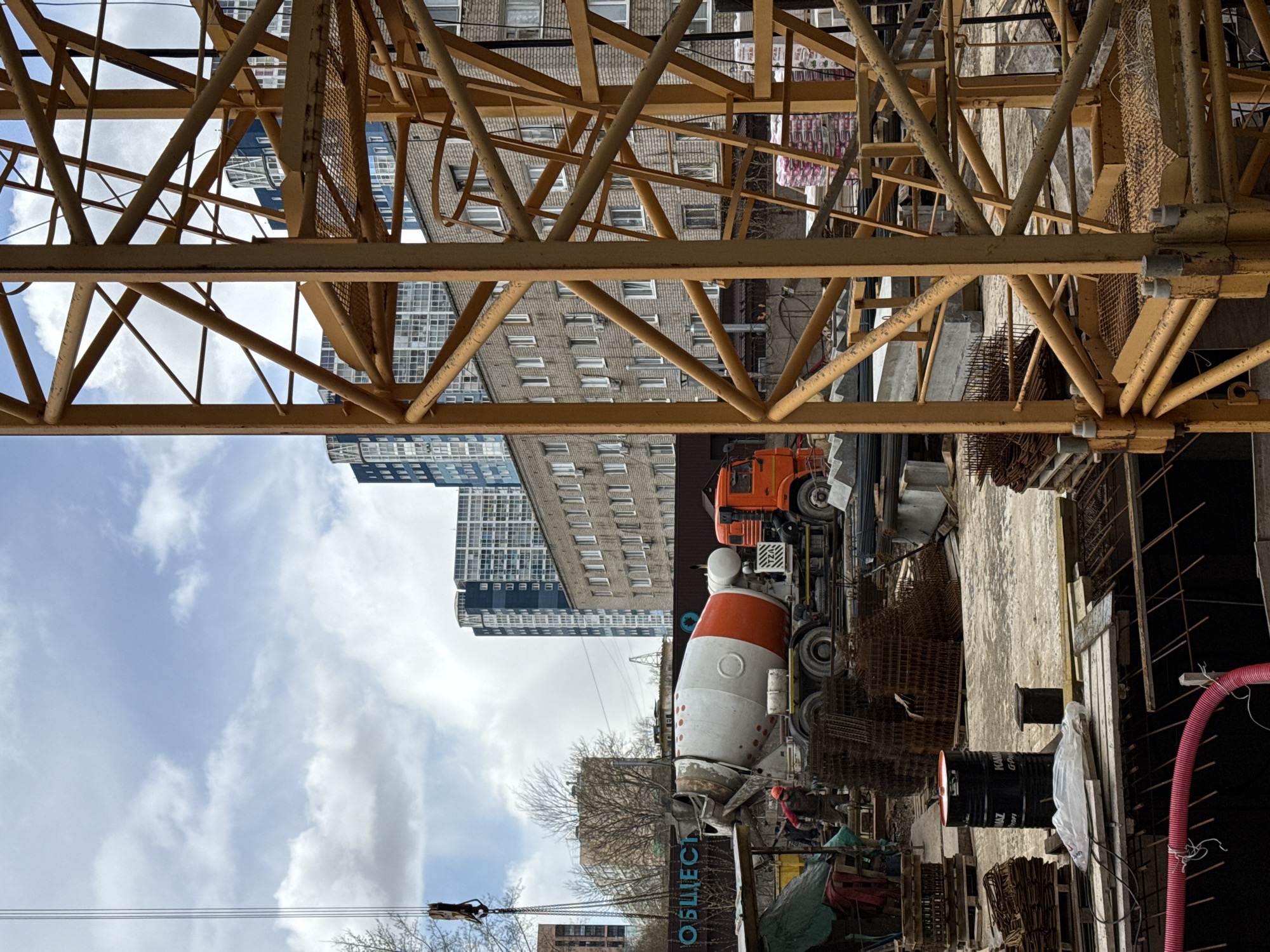 The image captures a dynamic construction scene in Perm, Russia, viewed through the intricate lattice framework of a large yellow crane or scaffolding structure. The foreground is dominated by the yellow metal beams, forming geometric patterns against the sky and background buildings.
In the mid-ground, a concrete mixer truck with an orange cab and a distinctive white-and-orange drum is parked. A worker wearing an orange hard hat and dark clothing is visible near the truck, suggesting activity related to concrete pouring or preparation. Scattered around the construction site are various materials, including bundles of rebar, wooden pallets, and a black barrel with "KAMAZ" visible on its side. A red corrugated pipe and other construction debris are also present. On the side of the concrete mixer's drum, partially visible Cyrillic text appears to read "ОБЩЕСТ...".
The background features several tall urban buildings, some with a grey, textured facade and numerous windows, others with a darker, more modern glass-fronted appearance. The sky above is partly cloudy with patches of bright blue. Bare trees line the distant horizon, indicating it might be late autumn or early spring. The scene is illuminated by daylight, suggesting it's daytime with clear weather. The overall impression is one of ongoing urban development and construction work.