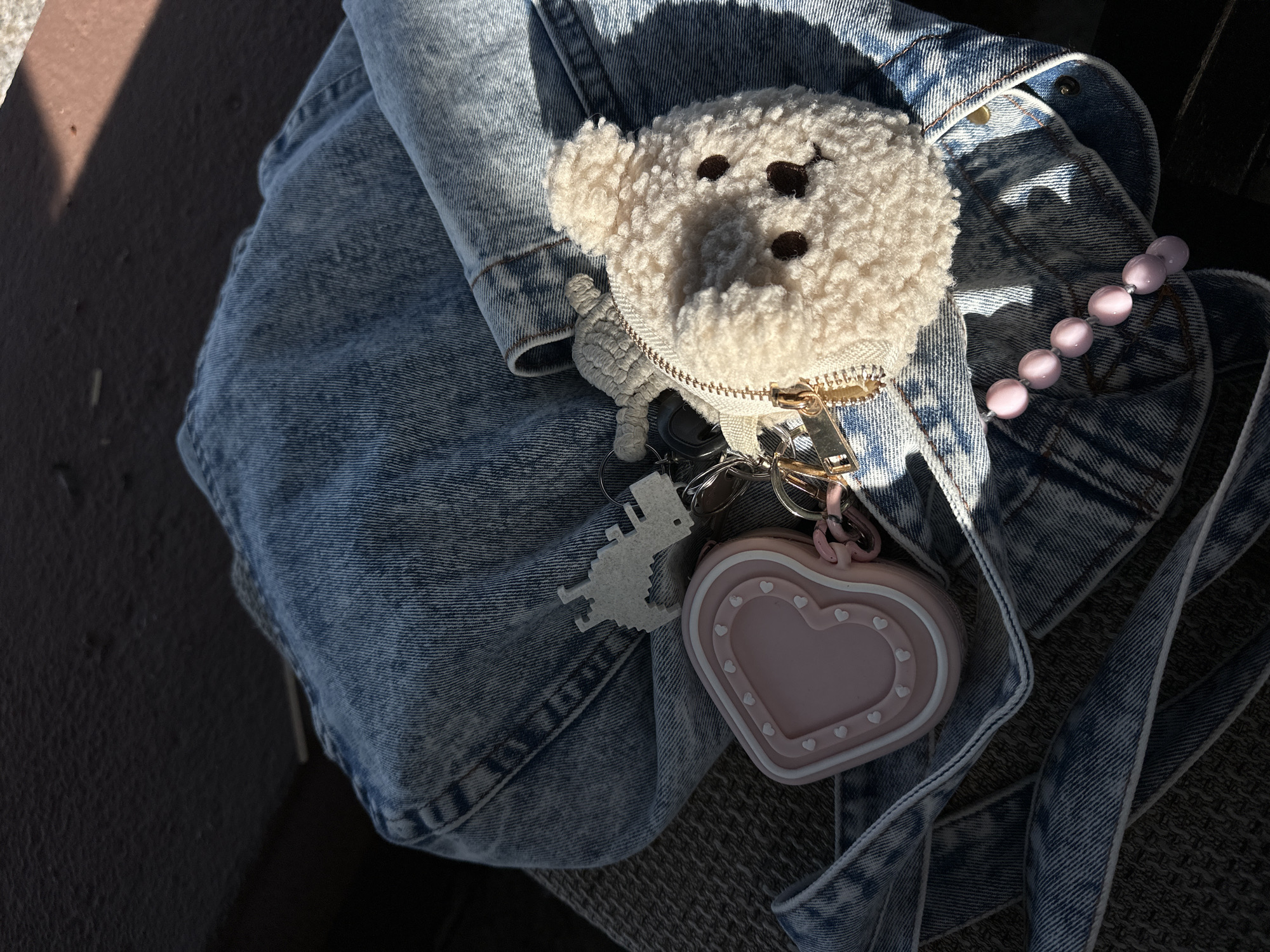 The image displays a close-up, top-down view of various personal accessories arranged casually on a surface. A light-wash denim fabric, likely part of a jacket, jeans, or a bag, forms the main backdrop for the items. Centrally featured is a cream-colored, fluffy teddy bear-shaped pouch or keychain, with embroidered dark brown eyes and nose, and a gold zipper.
Attached to a set of keys or keyrings near the teddy bear are a pixelated light-colored dinosaur keychain and a pink heart-shaped keychain or small case. The heart-shaped item has raised white heart details on its surface. To the right of the teddy bear, a strand of approximately six light pink, opalescent oval beads is visible.
The scene is illuminated by strong, direct sunlight, casting sharp shadows that indicate it is daytime, possibly mid-morning or afternoon. Parts of a dark, textured surface, perhaps concrete or upholstery, are visible around the denim, suggesting an outdoor or sunlit indoor setting. No specific activity or event is taking place, and there is no visible text in the image. The collection of items suggests everyday personal belongings. While the location context is Šušanj, Montenegro, this is not discernible from the visual cues in the photograph.