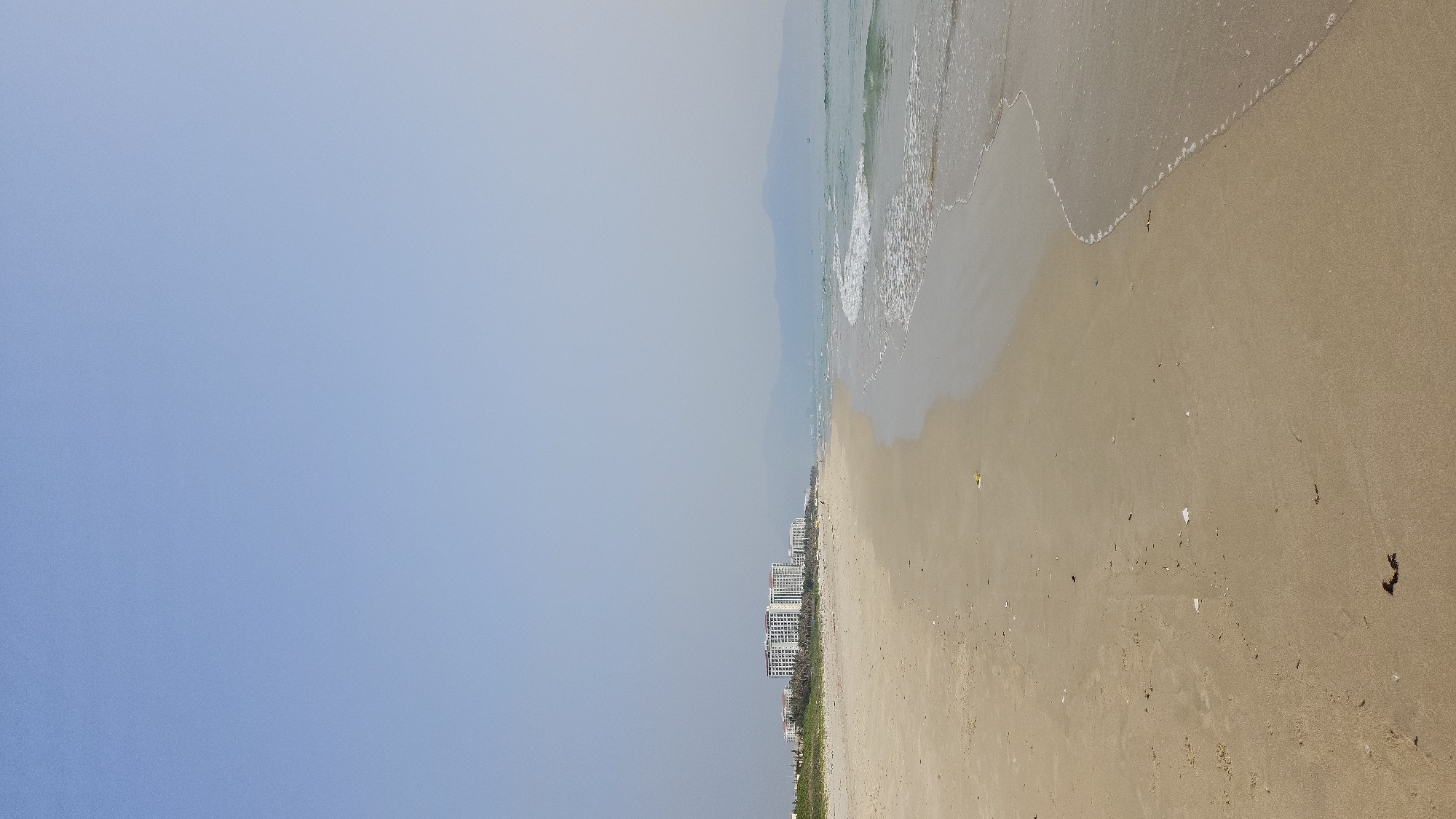 A wide, horizontal photograph captures a serene coastal scene in Bac Cam Ranh, Vietnam. The upper portion of the image is dominated by a vast, clear light blue sky, exhibiting a slight haze towards the distant horizon, suggesting a bright and sunny day. Below the sky, the calm ocean extends across the mid-ground, with gentle waves consistently breaking onto the shore, leaving foamy white patterns on the sand. The water itself appears a muted greenish-blue.
The lower half of the frame depicts a broad, light tan sandy beach. The sand shows natural texture and is sparsely scattered with small, dark pieces of debris, possibly seaweed, and other light-colored refuse, especially closer to the waterline. Along the horizon line, where the beach meets the land, a row of modern, multi-story white buildings, likely hotels or resorts, is visible, interspersed with green vegetation.
No people or specific activities are discernible, lending an atmosphere of tranquility and natural beauty to the scene. The image provides a peaceful view of a developed yet uncrowded beach destination during daylight hours. There is no visible text in the media file.