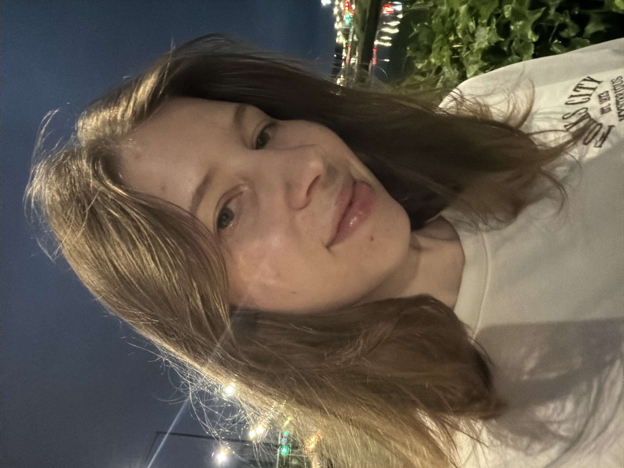A young woman with shoulder-length, light brown hair smiles towards the camera in a close-up selfie taken outdoors at night. She is wearing a white t-shirt with a graphic design. The background features blurry city lights, a body of water reflecting lights, and dark green foliage. The overall scene suggests an urban setting at dusk or night, possibly near a waterfront area within Dongducheon-si, South Korea.