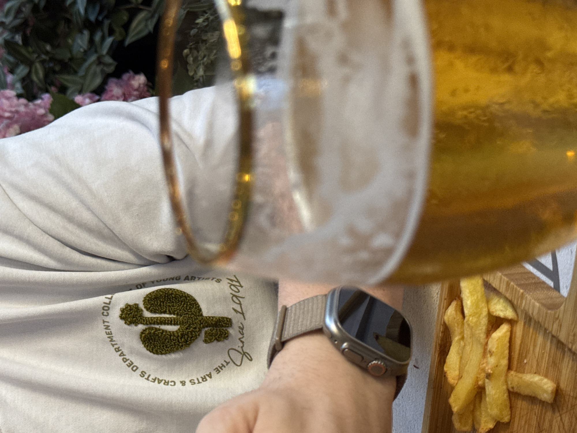 The image shows a close-up of a person's arm and chest, a glass of beer, and a plate of french fries. The person is wearing a light-colored sweatshirt with a circular logo featuring a cactus and text that reads "THE ARTS & CRAFTS DEPARTMENT COLL" and "OF YOUNG ARTISTS". Below the logo, the text "Since 1991" is visible. The person is also wearing a smartwatch. The glass of beer is partially in focus, showing a frothy head. The french fries are on a wooden board. The background is blurred and appears to be an outdoor setting with pink flowers and green foliage. There is no discernible location in the city of Plovdiv, Bulgaria.