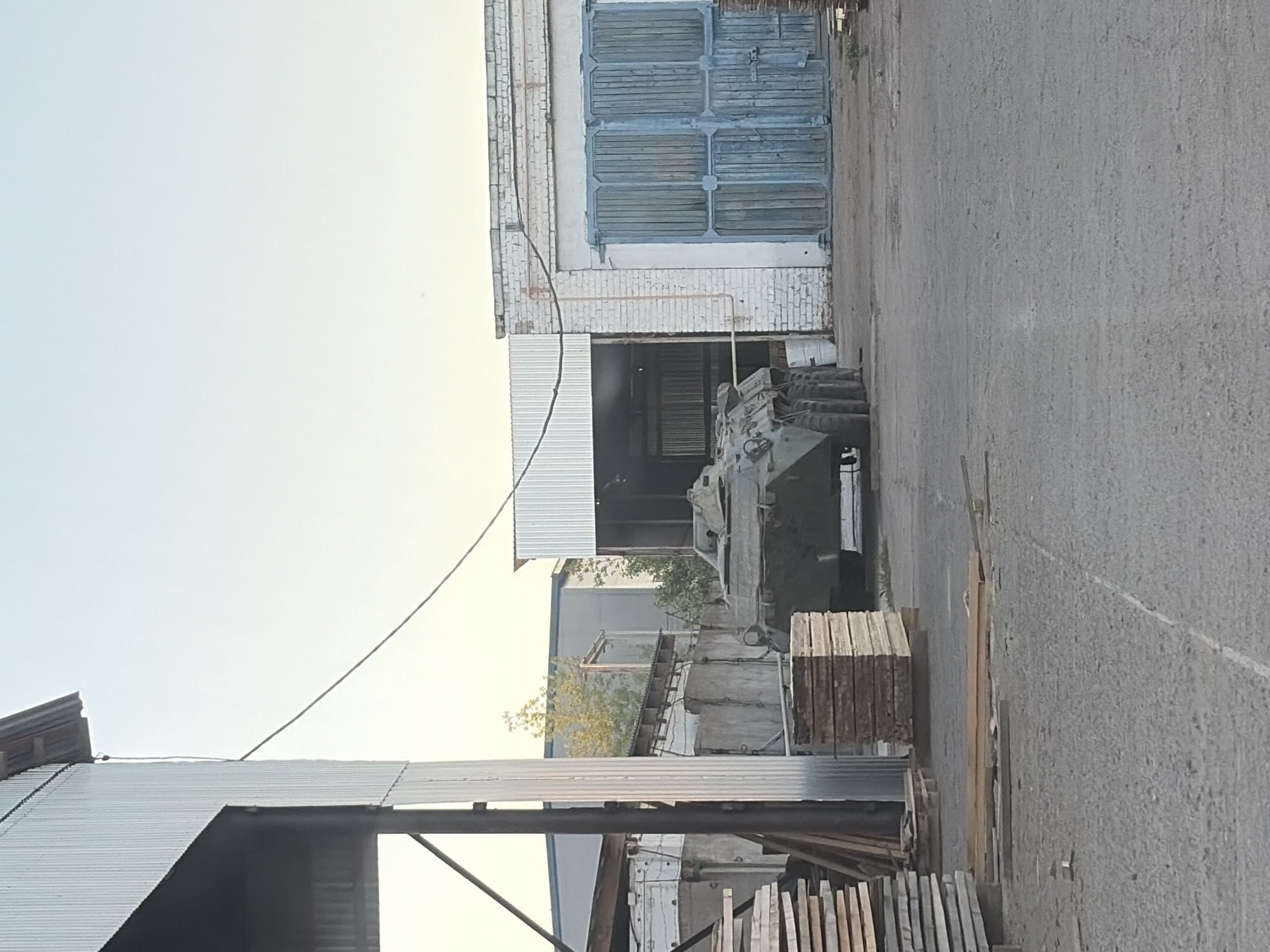 The image shows a military vehicle parked outside a building in what appears to be an industrial area. The building has a large, blue-painted door and a smaller opening where the vehicle is partially obscured. Stacks of lumber are visible in the foreground. The image does not contain any information about the city of Chaikovsky, Russia, and there are no people visible.