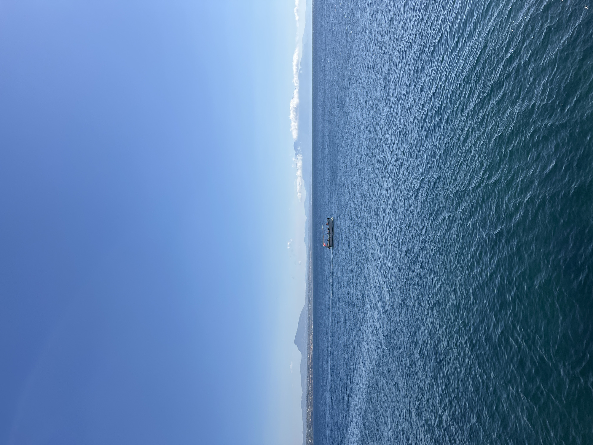 An image captures a wide expanse of the Mediterranean Sea off the coast of Terrasini, Italy. The water is a deep blue, with gentle ripples and waves. In the distance, a hazy shoreline with mountains is visible under a clear, bright blue sky. A small boat with several people on board is sailing on the sea. The boat has a flag with red and white stripes. The scene suggests a clear, sunny day suitable for sailing.