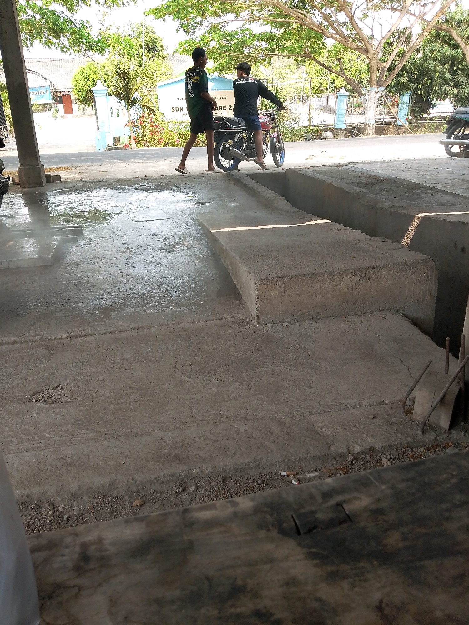 This image depicts an outdoor scene in the city of Kalitidu, Indonesia. In the foreground, a wet concrete floor with a trench is visible. Two young men are present. One man, wearing a black t-shirt and shorts, is sitting on a red motorcycle, holding its handlebars. The other man, wearing a green and black t-shirt and shorts, stands to the left of the motorcycle, facing away from the viewer. Both men are barefoot. In the background, a building with the sign "SDN 1 TENGGORO" is visible, along with trees and other structures. The overall setting appears to be an open area, possibly a workshop or a service area, indicated by the presence of the motorcycle and the wet concrete. The lighting suggests it is daytime, with sunlight casting shadows.
