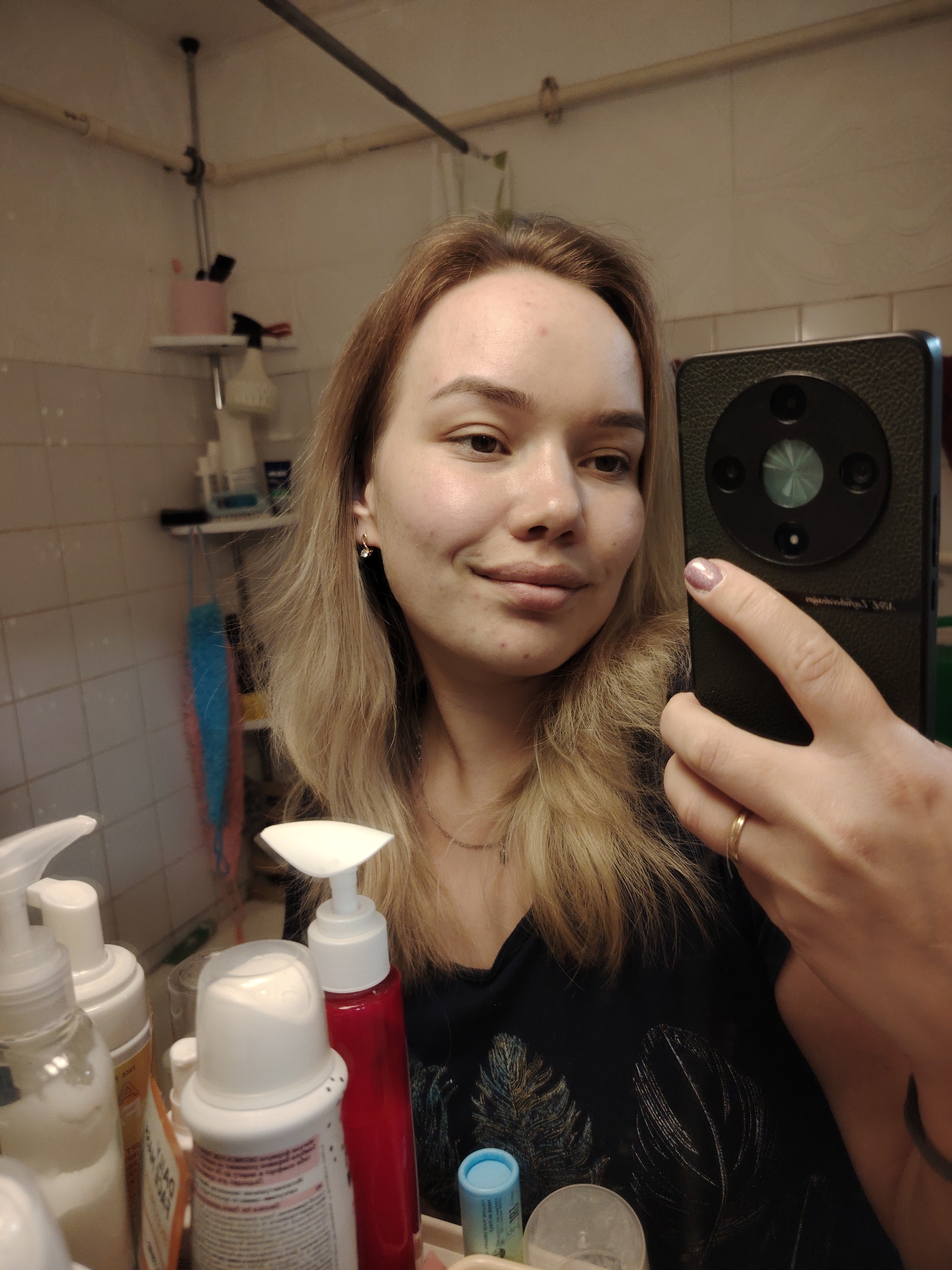 A person with blonde hair is taking a selfie in a bathroom. The person is holding a smartphone in their right hand, obscuring part of their face. The person is wearing a black t-shirt with a feather pattern. In the background are bathroom shelves with various personal care products. The bathroom has tiled walls. The location is Марковский, Russia.