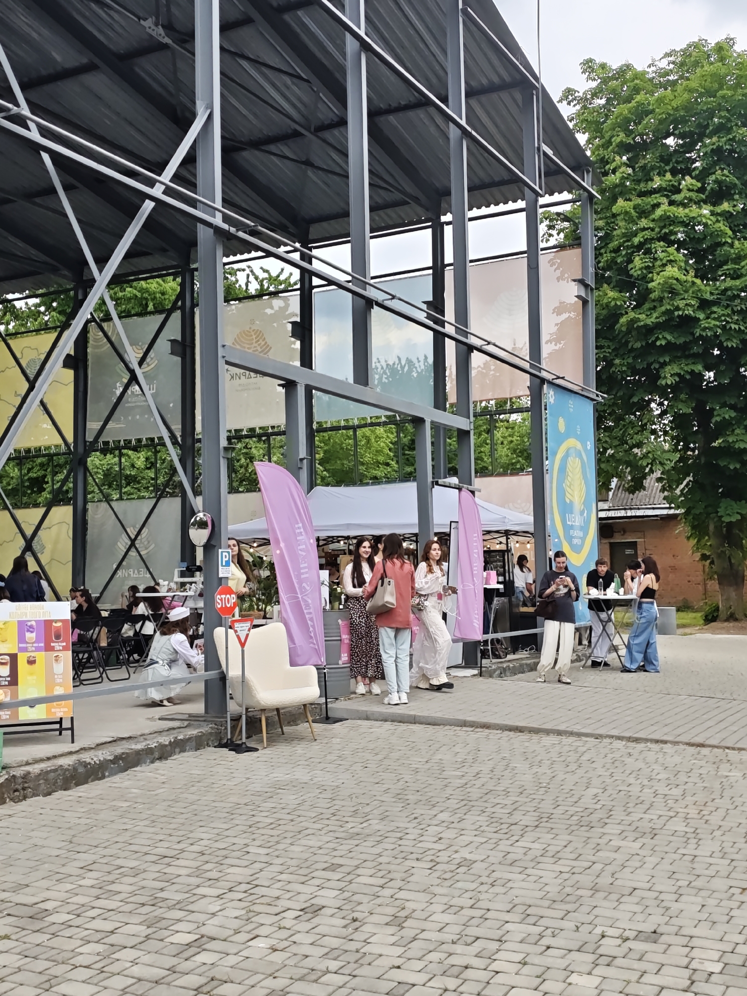 The image depicts an outdoor event taking place at "ЦЕДИК. Резиденція природи" (TSEDIK. Nature Residence) in Vinnytsia, Ukraine. The scene is dominated by a large, open-sided structural framework made of grey metal beams and a corrugated metal roof, providing a semi-covered space. Within this framework, multiple banners are suspended, including yellow ones with Cyrillic text such as "БЕЛЯРНК," and a prominent blue banner on the right featuring a stylized golden wheat stalk logo and the text "ЦЕДИК. РЕЗИДЕНЦІЯ ПРИРОДИ."
The ground is covered with grey rectangular paving stones. A concrete curb separates the main paved area from a lower, rougher concrete edge. Various people, primarily women, are present and engaged in different activities. On the far left, a promotional stand displays a "COFFEE MENU" with images of drinks and prices listed in Ukrainian Hryvnia (UAH). People are seated at black tables under the structure on the left. Near the center, a woman in a white uniform is kneeling on the ground.
Several individuals are standing in the central area, conversing. One woman is distinct in a red long-sleeved top and light blue wide-leg jeans. A pink vertical flag-banner with Cyrillic text ("ЦЕЛІНА") stands near a white armless upholstered chair. A red circular "STOP" sign and a red triangular sign are attached to a pole. To the right, outside the main structure, a group of three (two women and one man) are gathered around a small, round, foldable table. White canopy tents, indicative of vendor stalls, are visible in the background. A large deciduous tree with dense green foliage stands on the far right, partially obscuring a brick building. The sky is overcast.