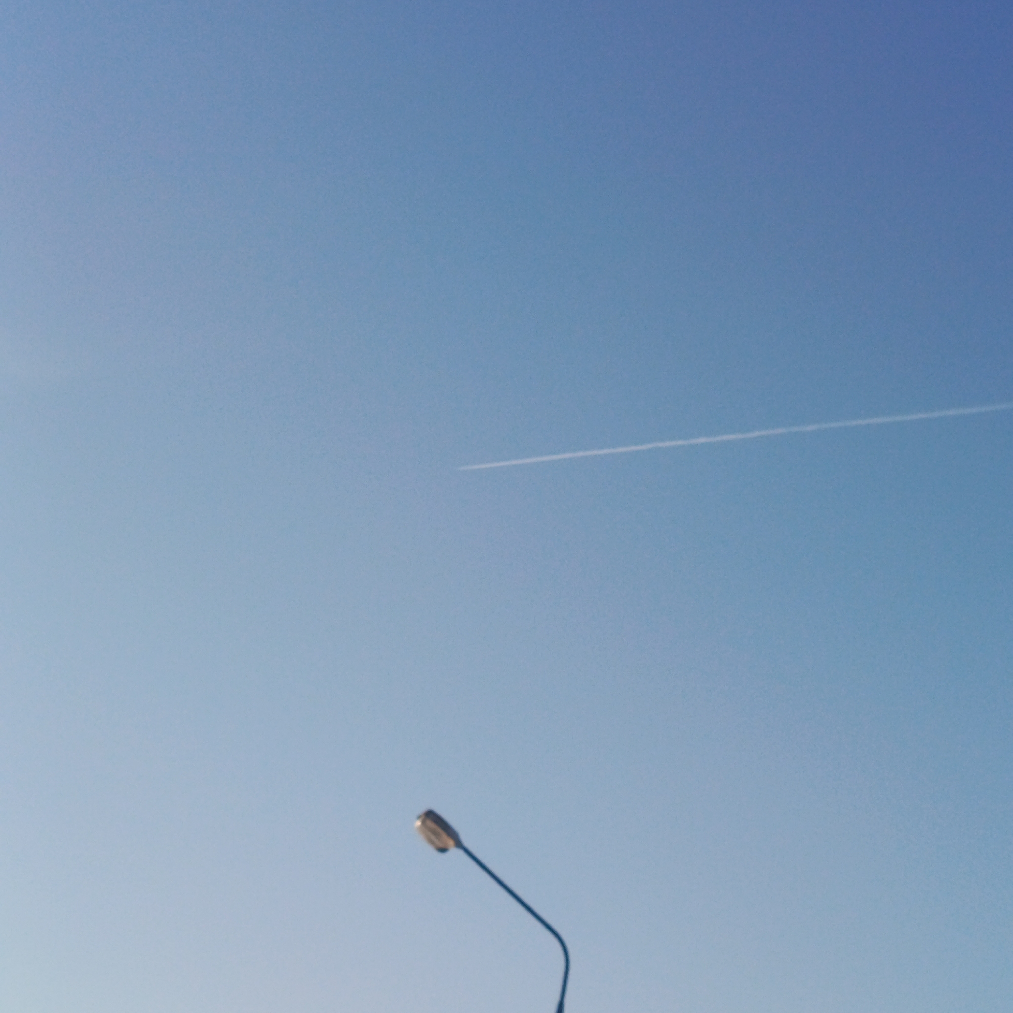 The image presents a clear blue sky with a faint contrail stretching across the upper right portion. A street lamp, with its bulb facing upwards, is positioned in the lower center of the frame. The overall environment suggests an outdoor setting, possibly a city. While the image includes information consistent with an outdoor setting in a city, it does not explicitly provide information establishing the location as Марковский, Russia.