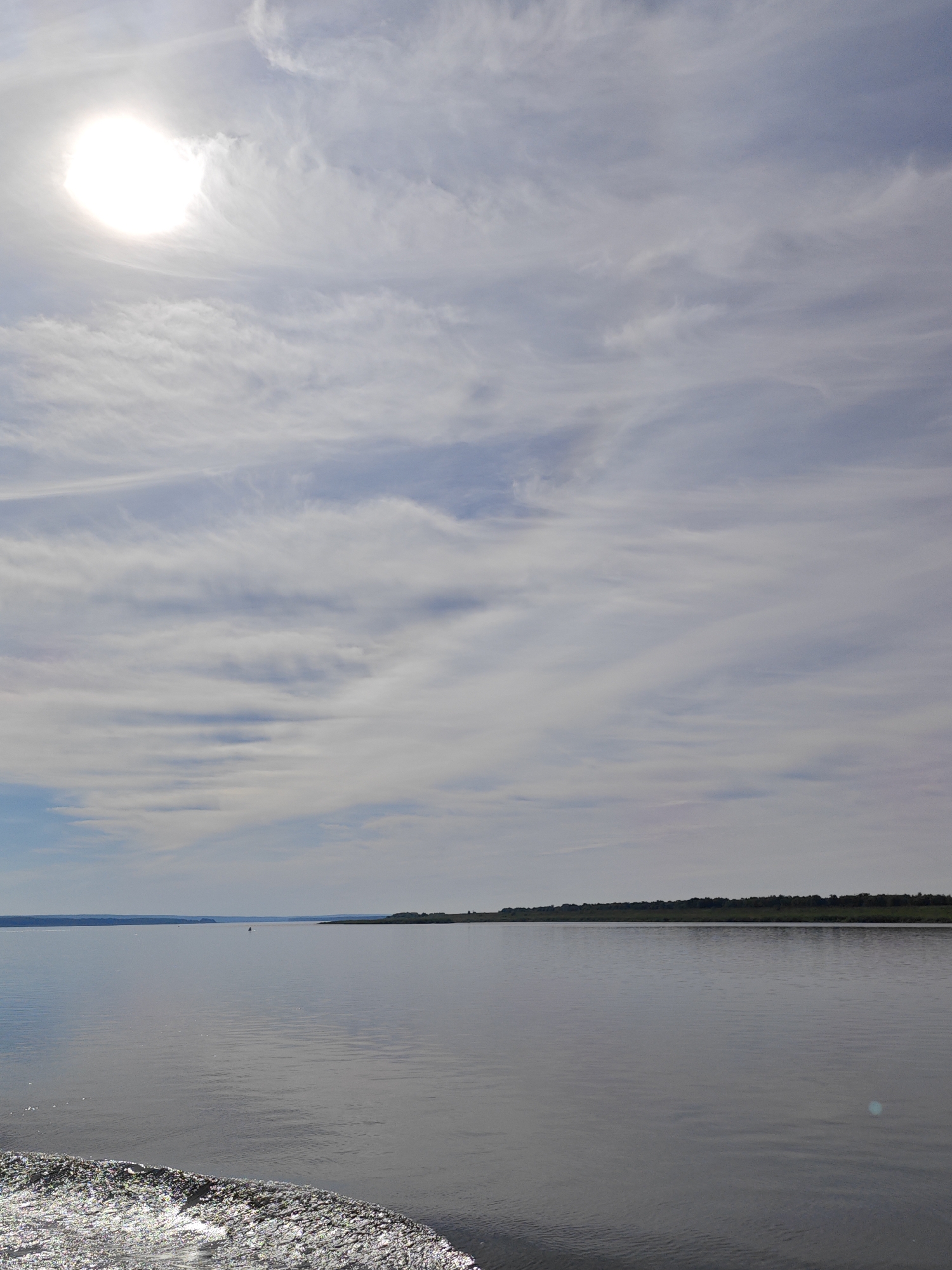 The image depicts a wide, calm body of water under a bright, partly cloudy sky. The sun is visible in the upper left corner, shining through wispy clouds that stretch across the sky in streaks. In the distance, a dark, tree-lined shore forms a horizontal line across the horizon. The water is smooth with a slight ripple effect in the foreground, suggesting movement or a gentle breeze. There is a small, dark object visible in the water in the middle distance, possibly a buoy or a small boat. The overall impression is one of a serene, natural landscape. The image is not geographically specific to Krivlyak, Russia.