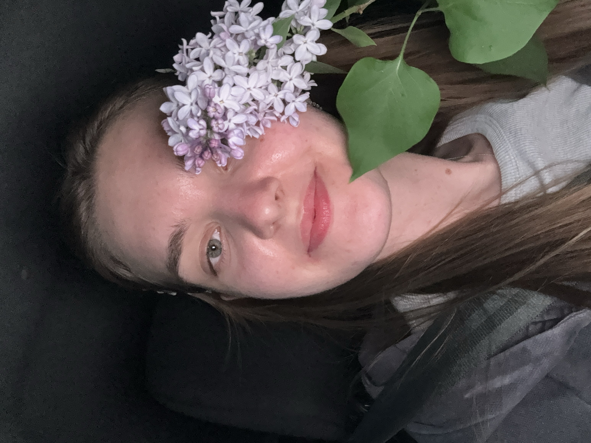 A woman with light brown hair is seen in an indoor setting, possibly a vehicle based on the gray material visible in the background. She is positioned at an angle with her face visible, adorned with lilac flowers placed near her forehead. A large green leaf is positioned near her cheek. Her expression is serene, with her eyes open. The overall image is well-lit and appears to be a casual snapshot. There is no evidence to suggest the image was taken in the city of Perm, Russia.