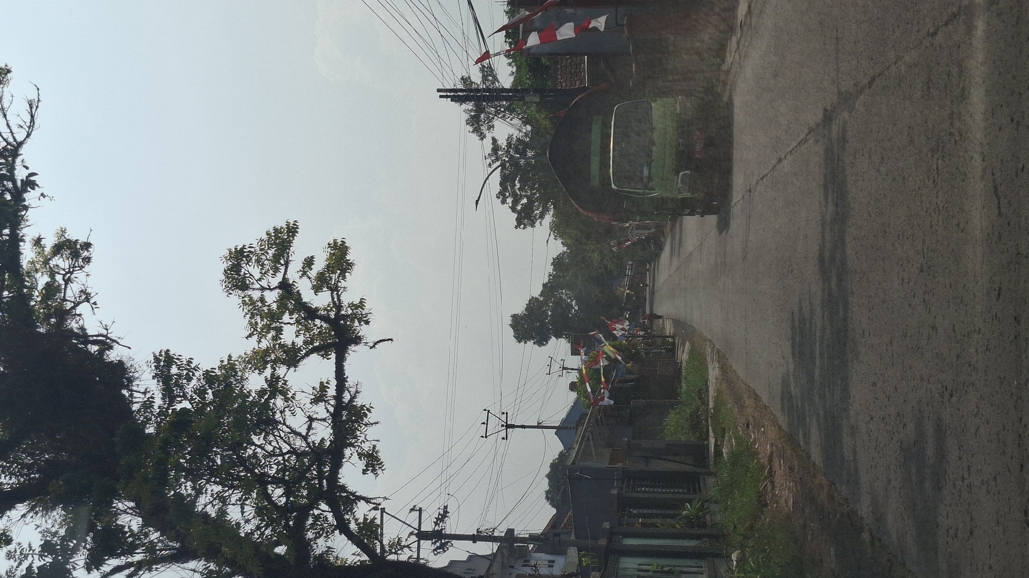 A vertical, eye-level, medium shot captures a street scene in Sukasari, Indonesia, on an overcast day. The road, paved with asphalt and showing visible cracks and patches, stretches into the distance, flanked by houses and vegetation. In the foreground, a large tree with dense foliage and mossy branches partially obscures the upper left quadrant of the frame, casting shadows on the road.
A green truck with a covered cargo bed is the most prominent vehicle, traveling away from the viewer down the right side of the road. Its license plate is partially visible, displaying characters that appear to be "F 0056 U." A single pedestrian, wearing red clothing, walks in the distance on the left side of the road.
Along the sides of the street, rows of houses with dark roofs and muted-colored walls are visible. Indonesian flags, red and white, are hung along the route, indicating a possible national celebration or event. Power lines crisscross the sky, supported by utility poles. The environment appears to be a residential area with a somewhat rural character, characterized by abundant trees and vegetation. The sky is a pale blue, suggesting hazy or cloudy conditions.