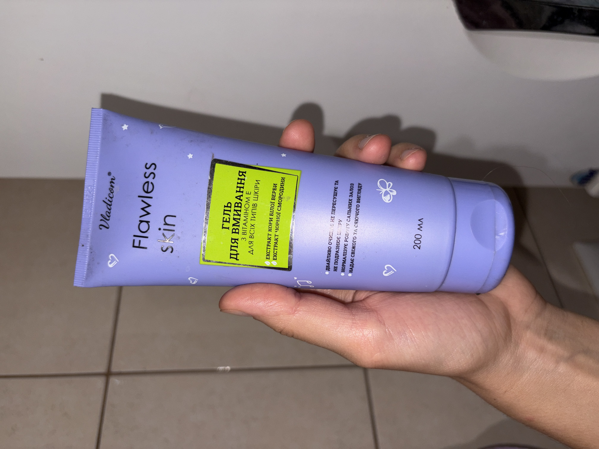 A person's hand holds a light purple tube of "Flawless Skin" facial cleanser. The tube has a lime green label with black Ukrainian text. The text indicates the product is a gel for washing with Vitamin E for all skin types, and contains white birch bark extract and blackcurrant extract. It also states that it gently cleanses without drying or irritating the skin, normalizes sebum production, and gives a fresh look. The volume is listed as 200 ml. The background appears to be a tiled wall in a bathroom.