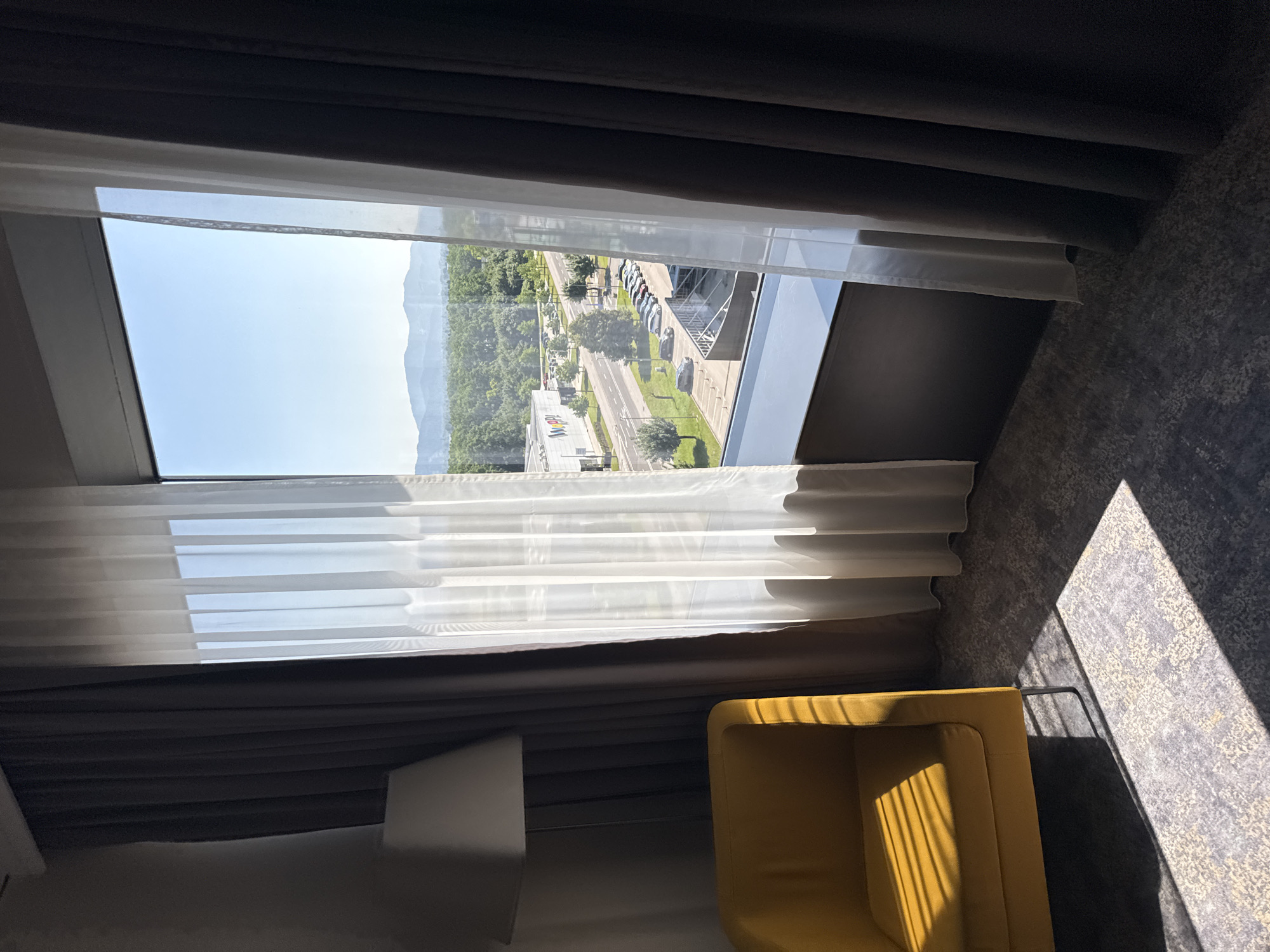 The image captures a hotel room interior looking out through a large window. In the foreground, a bright yellow armchair is positioned on a patterned gray carpet. Sunlight casts distinct striped shadows from what appears to be window blinds onto the armchair and the carpet. To the left, a floor lamp is partially visible behind the armchair and dark curtains. The window itself is covered by sheer white curtains and dark drapes.
Beyond the window, a cityscape unfolds under a clear blue sky. A road with parked cars and a few trees borders a modern building with signage that reads "WOPPI". In the distance, lush green trees form a dense forest, leading to rolling hills and mountains that frame the horizon.
The image appears to have been taken in Ljubljana, Slovenia, based on the architectural style of the building and the presence of the "WOPPI" signage, which likely corresponds to a commercial establishment in the city. The overall scene suggests a well-lit hotel room with a view of the outskirts of Ljubljana, possibly in an area with commercial and natural landscapes. There are no people or animals visible in the frame, nor any discernible actions or interactions occurring. The image is a static, observational snapshot of the room and its view.