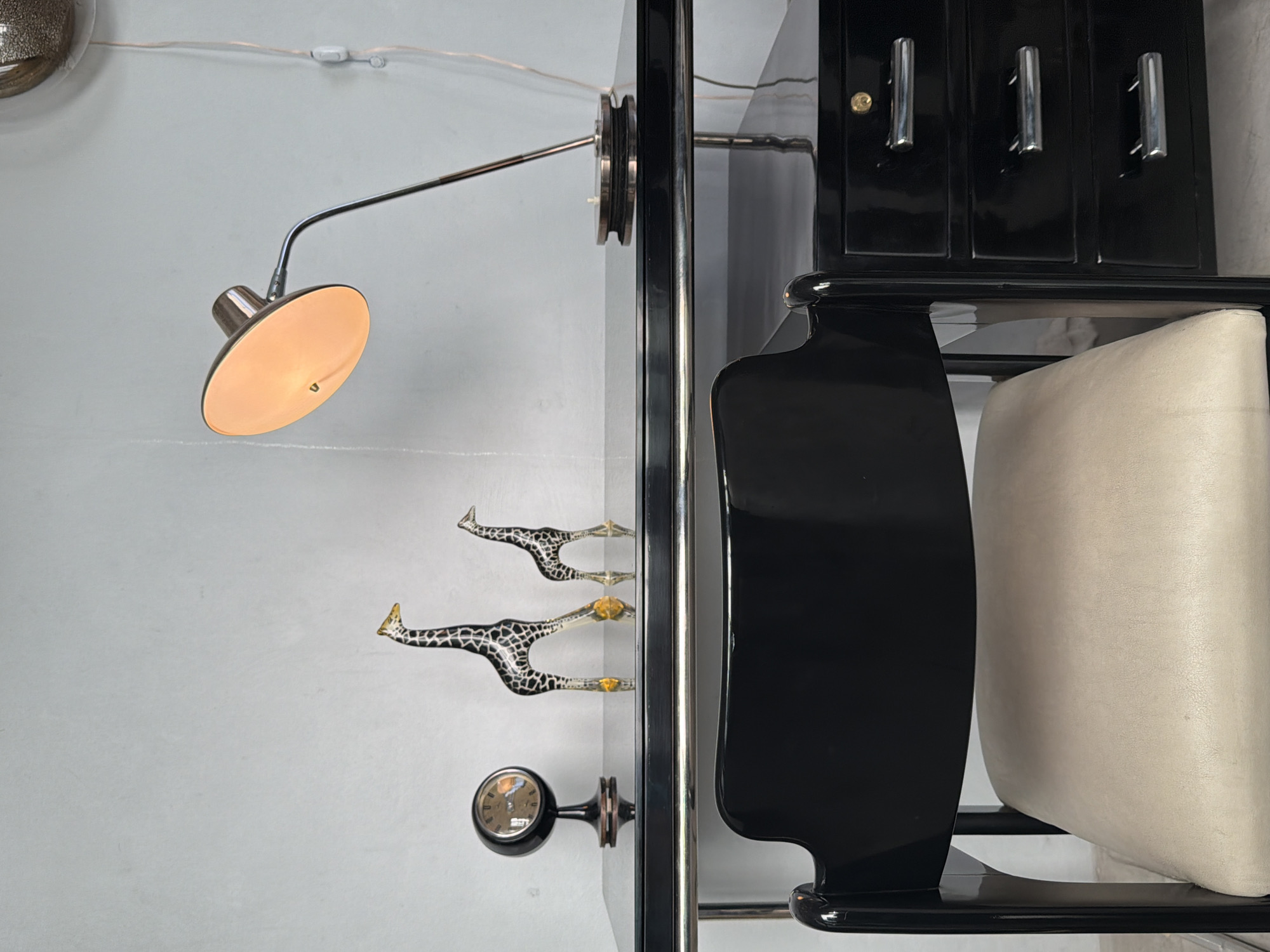 This media file captures an indoor scene, likely a room or display area, from a low, vertical perspective. The main subjects are various wall-mounted items and a sleek furniture piece.
On the left side, a light-colored wall serves as the background. In the upper left corner, a partial view of a metallic, reflective, circular object is visible. Below it, a modern wall-mounted lamp extends with a metallic arm, ending in an illuminated, shallow, light-colored conical shade. Further down the wall, three stylized decorative items resembling giraffes, featuring black and white spotted patterns and gold-colored details, are mounted vertically, possibly functioning as hooks. Below these, a small, round clock with Roman numerals is also affixed to the wall. Thin electrical wires run along the upper part of the wall.
The right side of the image is dominated by a glossy black furniture piece, possibly a cabinet or part of a vanity unit. Its upper section features three prominent chrome pull handles and a small brass-colored knob. Beneath this, a cream or beige upholstered cushion, likely the seat of a chair or stool, is tucked in, complementing the dark furniture. The furniture exhibits smooth, curved lines.
There is no activity taking place, and the scene is static. The lamp being lit suggests it might be evening or an intentionally lit space. The overall aesthetic, with the glossy black furniture and chrome accents, hints at a modern or Art Deco design. No specific text is clearly legible on the clock face beyond Roman numerals. The location context provided is Buenos Aires, Argentina, though no direct visual cues within the image confirm this.