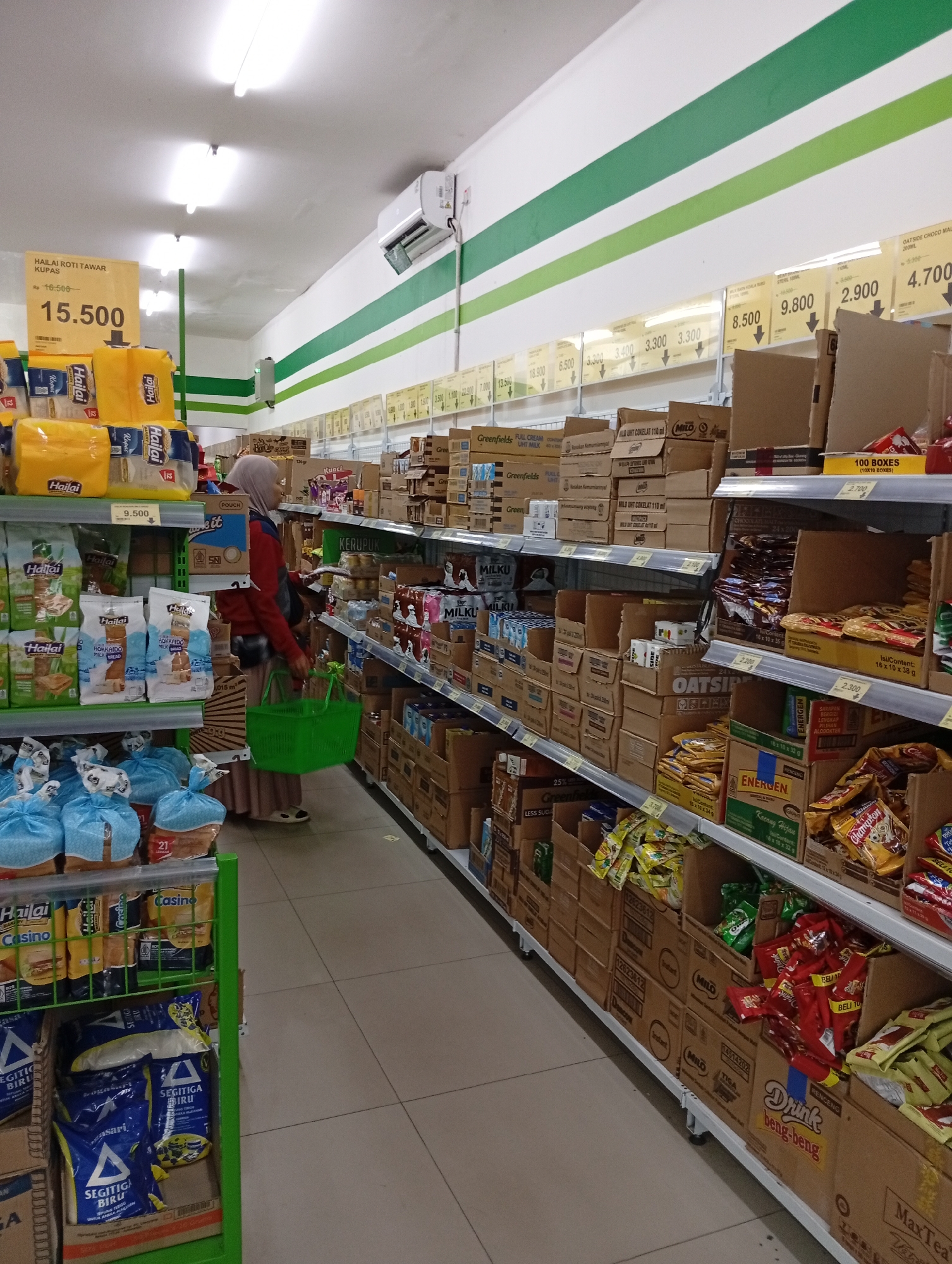 This is a medium-shot, eye-level, indoor photograph of a grocery store aisle in Babakan, Indonesia. The aisle is filled with shelves stacked high with various food products, primarily packaged goods and boxes. On the left side of the frame, a woman wearing a hijab and a red top is browsing the shelves. She is holding a bright green shopping basket in her right hand. The shelves are well-stocked with items like bread, milk cartons, and bagged snacks. Prominently displayed are yellow bags of "Hailai Roti Tawar Kupas" with a price tag of 15,500.
The right side of the aisle features more shelves laden with stacked boxes and packaged items. There are various brands and types of products visible, including what appear to be cereal boxes, snacks, and drinks. Some price tags are visible on the shelves, such as 2.900 and 4.700. The overall environment suggests a typical supermarket or minimart. The lighting is bright, likely from overhead fluorescent lights. The floor is tiled with a neutral color. The background shows more aisles and shelving, indicating a larger retail space. The image is clear and in focus, capturing the details of the products and the shopping activity.