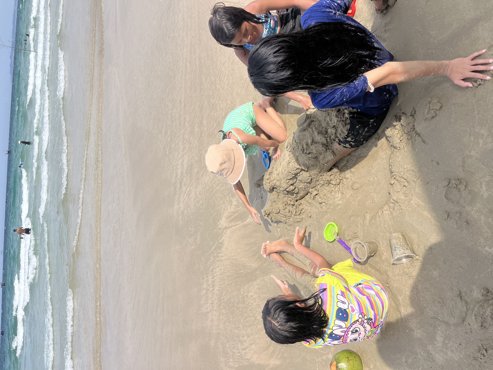 This is a medium-shot, eye-level, outdoor photograph taken on a beach in Nam Nha Trang Ward, Vietnam. The image depicts four children playing in the sand. In the foreground, a young girl with long black hair, wearing a yellow shirt with rainbow stripes and a cartoon character print, sits with her back to the camera, facing the ocean. She has wet sand on her legs and arms and is pointing towards a sandcastle. To her left are a green coconut and a purple shovel with a green sieve attachment, and two plastic cups, one containing wet sand.
Behind her, a second girl, facing away from the camera, is partially buried in sand, wearing a dark blue t-shirt and black shorts. Her arm is extended in front of her.
To the right of the sandcastle, a third child, wearing a beige sun hat and a green and white striped swimsuit, is kneeling and actively playing in the sand. A blue toy shovel is next to her.
Further to the right, a fourth child, with long black hair, is wearing a patterned blue and white swim top and black shorts. She is sitting and looking down at the sandcastle.
The sand is wet and uneven, with some footprints and the beginnings of a sandcastle. In the background, the ocean has small to medium waves, and the sky is bright and overcast. Several distant figures can be seen in the water and on the shore, and a few boats are visible on the horizon. The overall impression is a sunny day at the beach, with children engaged in sand play.