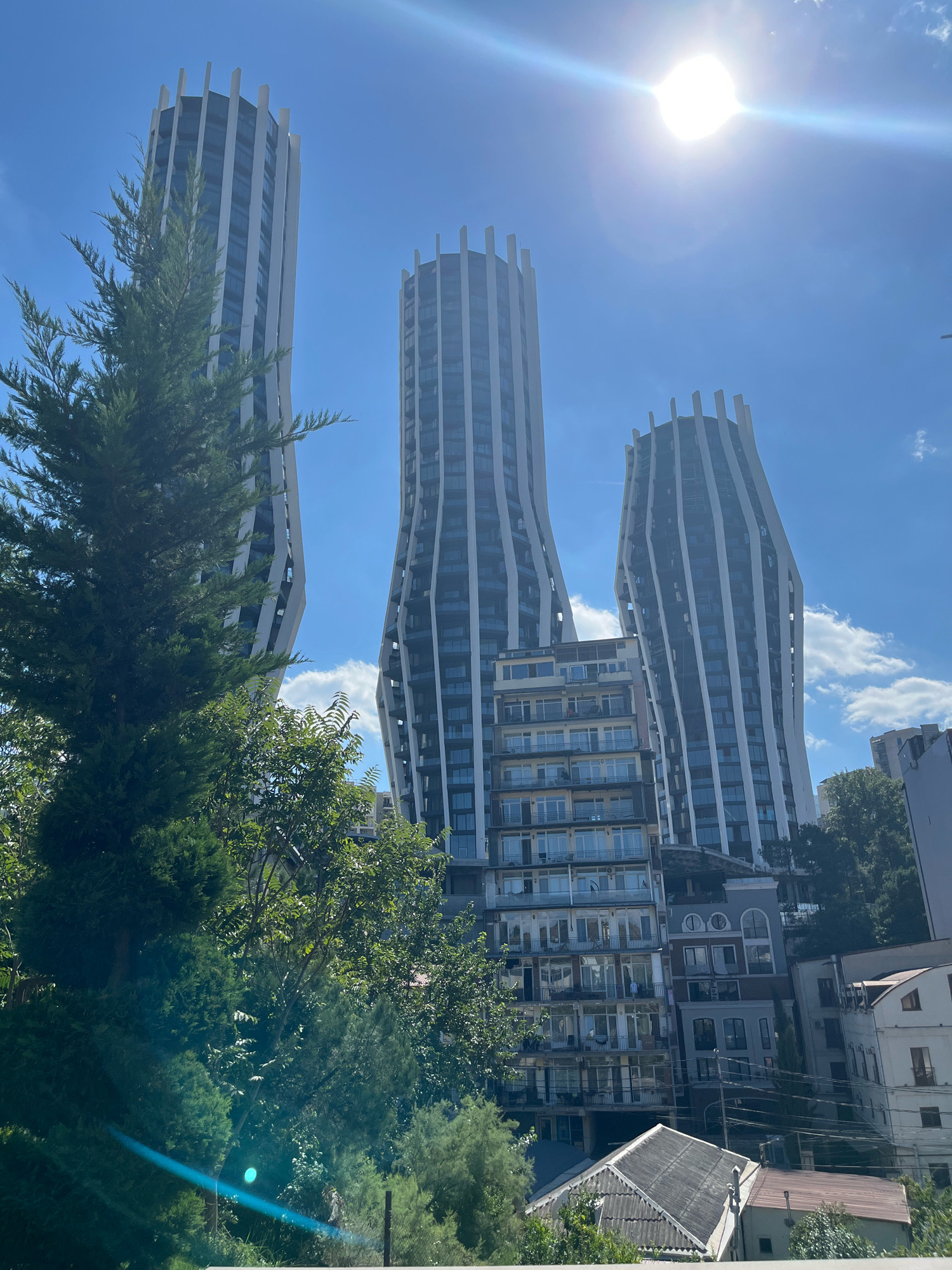 In the city of Tbilisi, Georgia, an outdoor shot features a bright, sunny day with a clear blue sky and scattered clouds. In the foreground, lush green trees and foliage dominate the left side of the frame. Behind the trees, three distinctive, tall, modern buildings rise into the sky. These buildings have a unique, undulating architectural design, with white vertical elements interspersed with dark glass windows, creating a wave-like appearance. The sun is prominently visible in the upper right quadrant of the image, casting a lens flare that creates a bright, starburst effect. Below the modern towers, several older, multi-story residential buildings with balconies and windows are visible, hinting at the diverse architectural styles present in the city. The overall scene depicts a juxtaposition of modern and older urban architecture against a backdrop of natural greenery and bright sunlight.