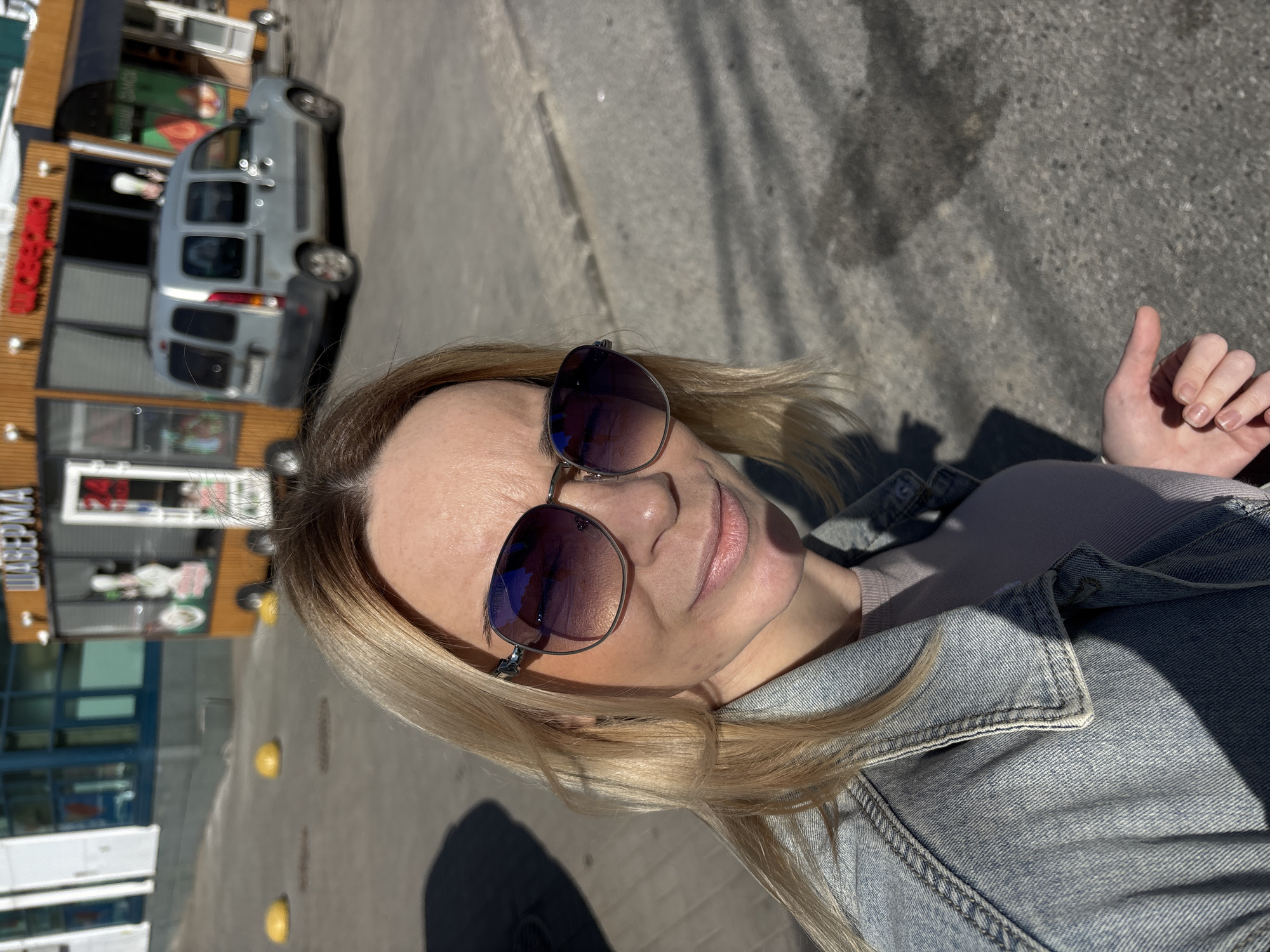 A rotated selfie captures a blonde-haired woman outdoors on a sunny day. She is wearing dark sunglasses with reflective lenses and a light blue denim jacket over a lighter-toned top. Her left hand is visible, giving a thumbs-up gesture.
The background shows an urban environment. To her left, behind her shoulder, is a light grey commercial van, possibly a Renault Kangoo, parked near a building. This building features a wooden facade and glass windows, typical of commercial establishments. Partially visible text in Cyrillic script on the building's facade reads "ШАУРМА," indicating a fast-food or kebab shop.
The ground is paved with asphalt or concrete, and several yellow concrete bollards are visible. Strong, defined shadows cast by the subjects and objects suggest bright daylight, likely midday or early afternoon, with clear weather conditions. The reflections in the woman's sunglasses hint at the bright sky and surrounding environment. The image has been rotated counter-clockwise from its original orientation.