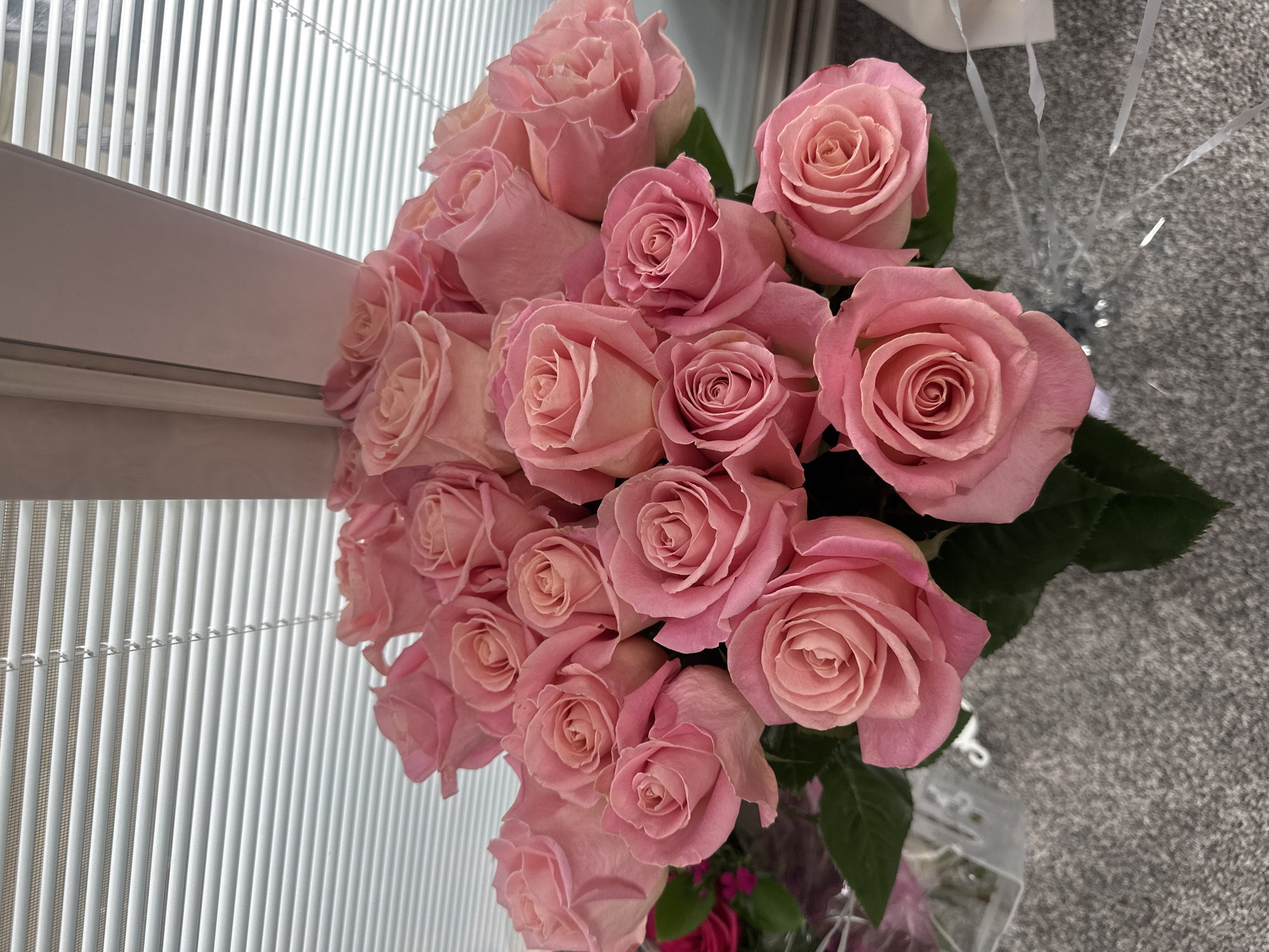 A bouquet of pink roses is displayed against a background of white blinds and a light pink wall. The roses are various shades of pink, with some lighter and some darker. They are arranged in a dense cluster, with some roses fully bloomed and others still in bud. The bouquet is placed on a gray, textured surface, possibly a carpet or a table runner. There are some hints of green leaves among the roses. The lighting suggests an indoor setting, possibly near a window. The image appears to be a close-up shot, focusing primarily on the flowers. There is no indication of the location being in Schaumburg, United States of America.