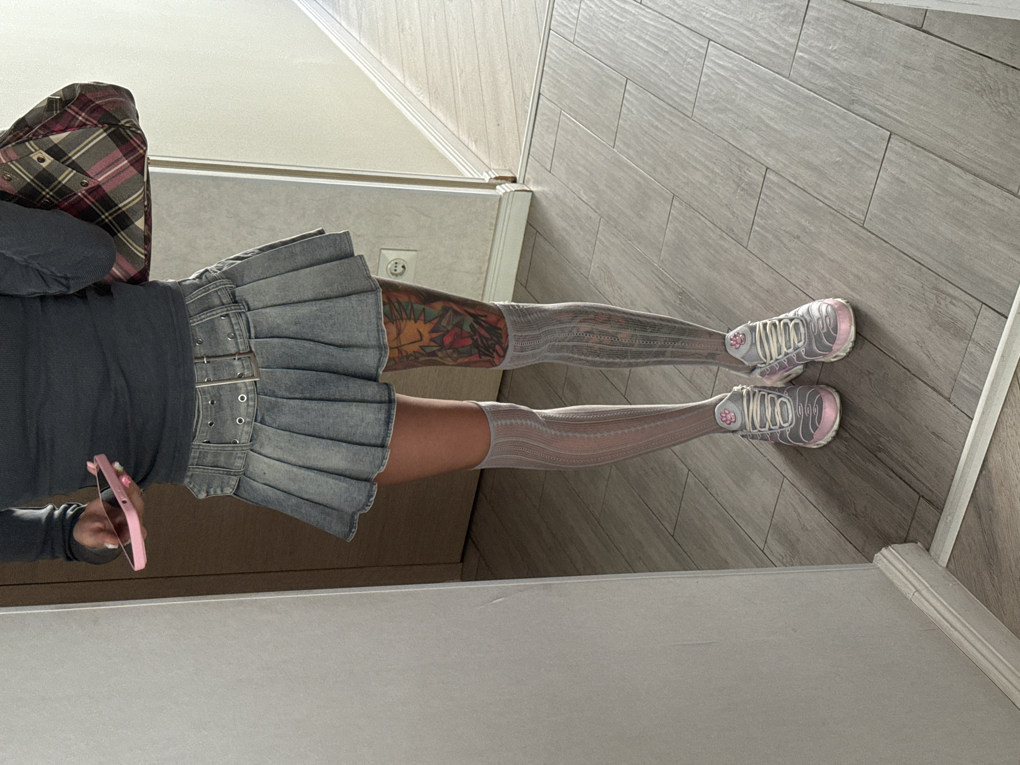 A person stands in a hallway, viewed from the waist down, holding a pink smartphone. They are wearing a grey long-sleeved top, a light blue denim pleated mini-skirt with a double-buckle belt, and white and grey patterned knee-high socks. Visible on their right leg is a colorful tattoo. They are wearing grey and pink athletic shoes. The floor is tiled with light grey wood-look planks. To the left is a white wall, and to the right is a doorway leading to a brightly lit area. The scene appears to be indoors, possibly in a residential setting. There is no indication that this location is in Moscow, Russia.