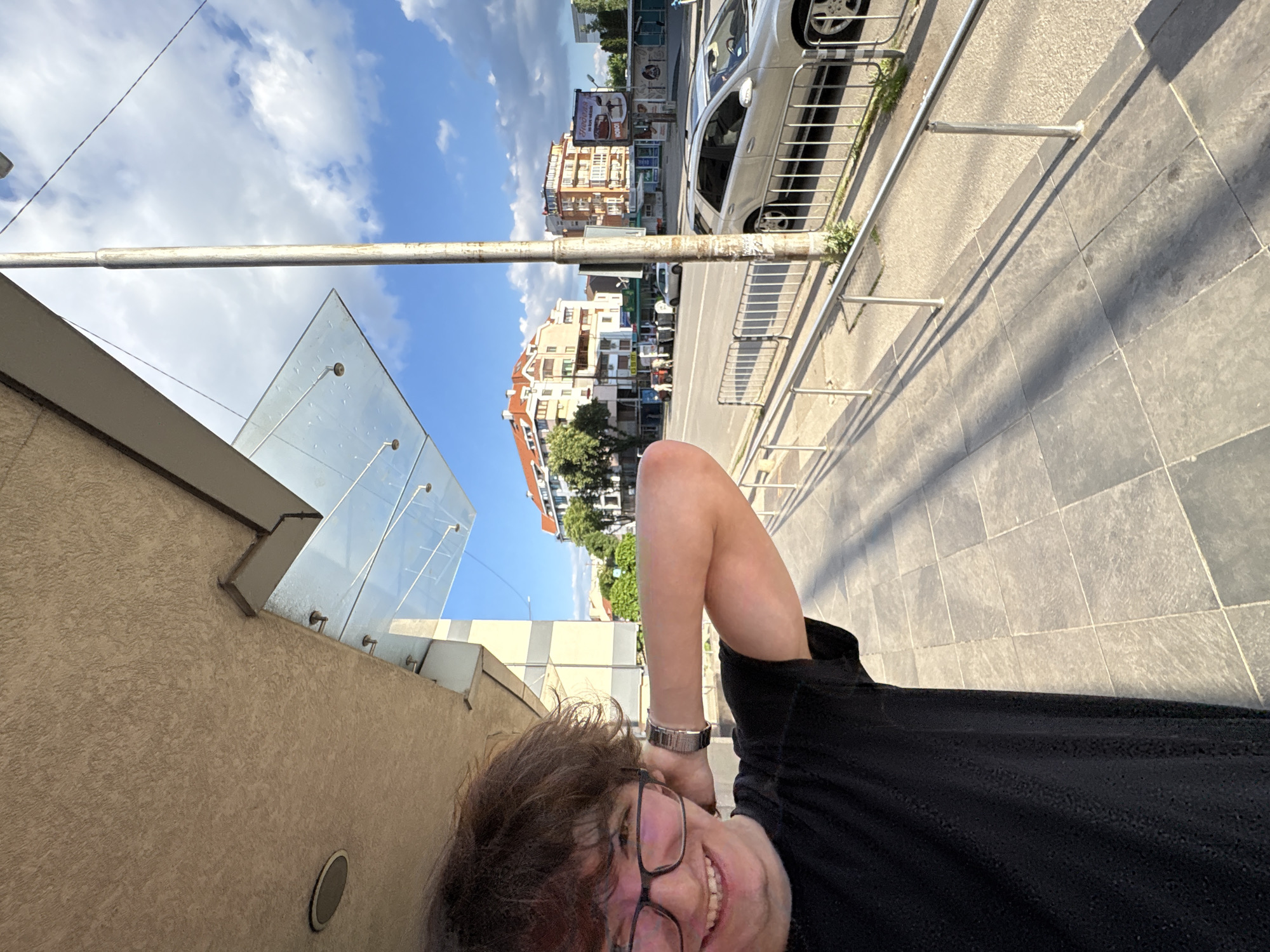 A selfie-style, tilted, high-angle, medium shot shows a person in the foreground and a street scene in Sofia, Bulgaria, in the background. The person is wearing glasses, a silver watch, and a black shirt. Their head is tilted to the left, and they are smiling slightly.
The background features a sidewalk made of large square pavers, a silver metal guardrail, and a street with cars and buildings. The buildings are a mix of residential and commercial structures, with varying architectural styles. The sky is blue with scattered white clouds. A building with a glass-covered entryway is partially visible on the left. The angle suggests the person is lying down or leaning back at an extreme angle while taking the photograph.