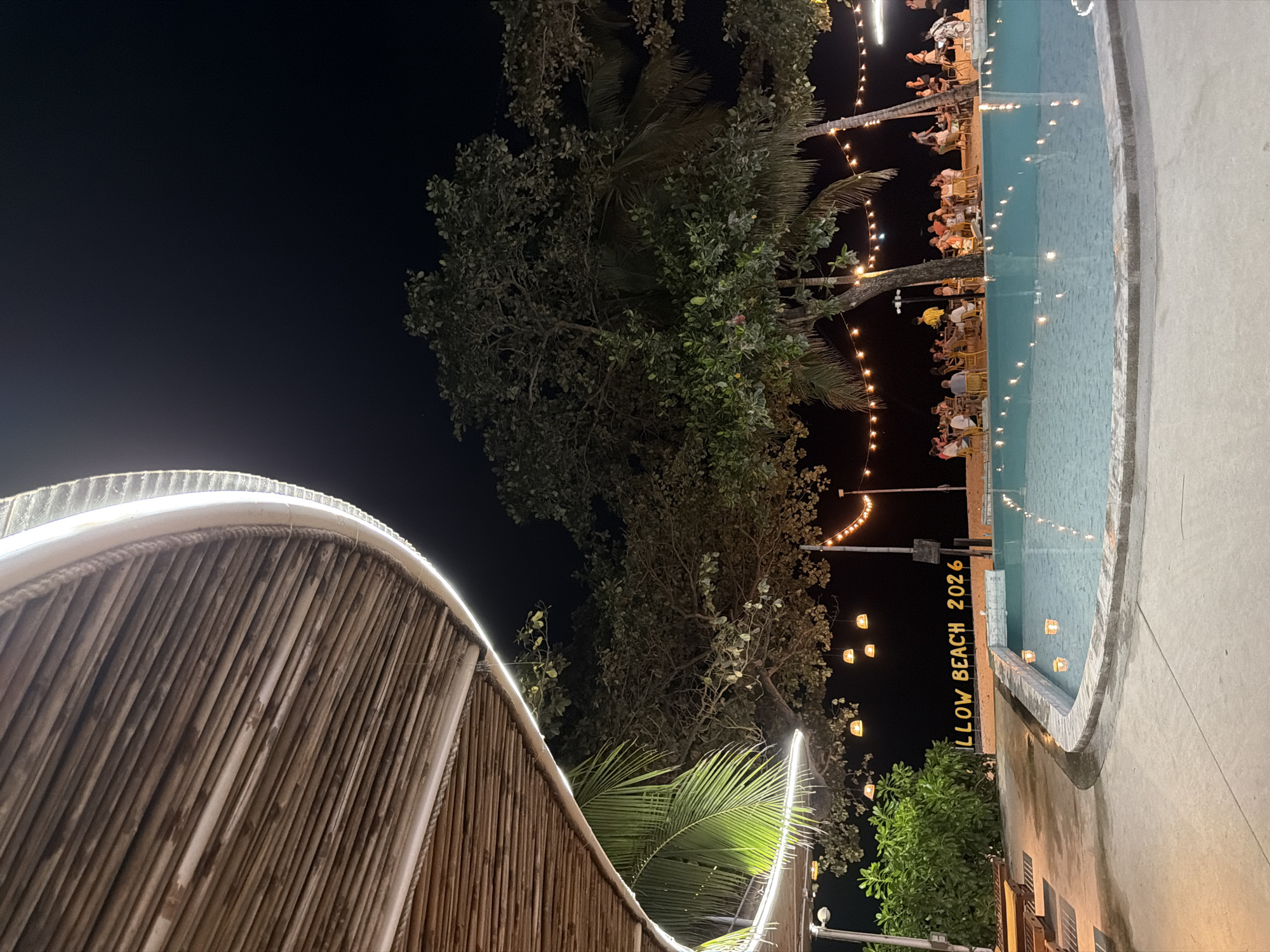 A nighttime scene at a beachside establishment in Ko Samui, Thailand, features a swimming pool in the foreground and a dining area in the background. The pool's edge is illuminated by floating lights, and the water reflects the surrounding string lights. Behind the pool, a sandy area is set up with tables and chairs where patrons are dining. Palm trees and other foliage are visible, with string lights draped between them, creating a festive atmosphere. In the distance, illuminated signage reads "LLOW BEACH 2026". The roof structure of the establishment, made of bamboo and adorned with LED strip lights, curves over the left side of the image.