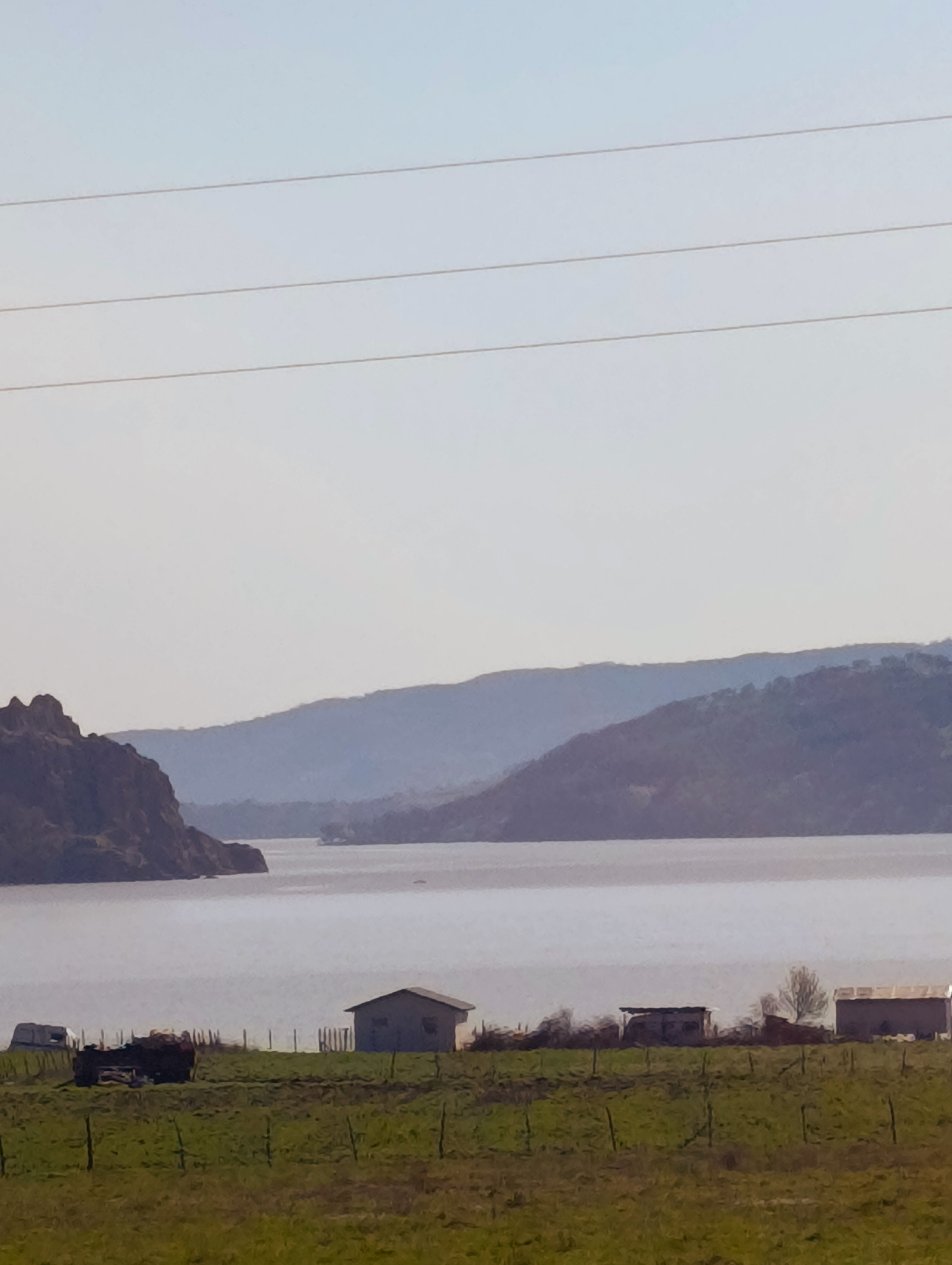 The image presents a landscape scene near Shiroko Pole, Bulgaria. In the foreground, a grassy field with a visible fence line dominates the lower portion of the frame. A collection of weathered buildings and structures, including what appears to be a trailer, a shed, and possibly a small barn, are situated along the edge of the field near the water. Further out, a body of water extends towards the horizon, flanked by hills or mountains on either side. The overall lighting suggests either dawn or dusk, casting a soft, muted tone across the scene. Power lines are visible in the upper part of the frame.