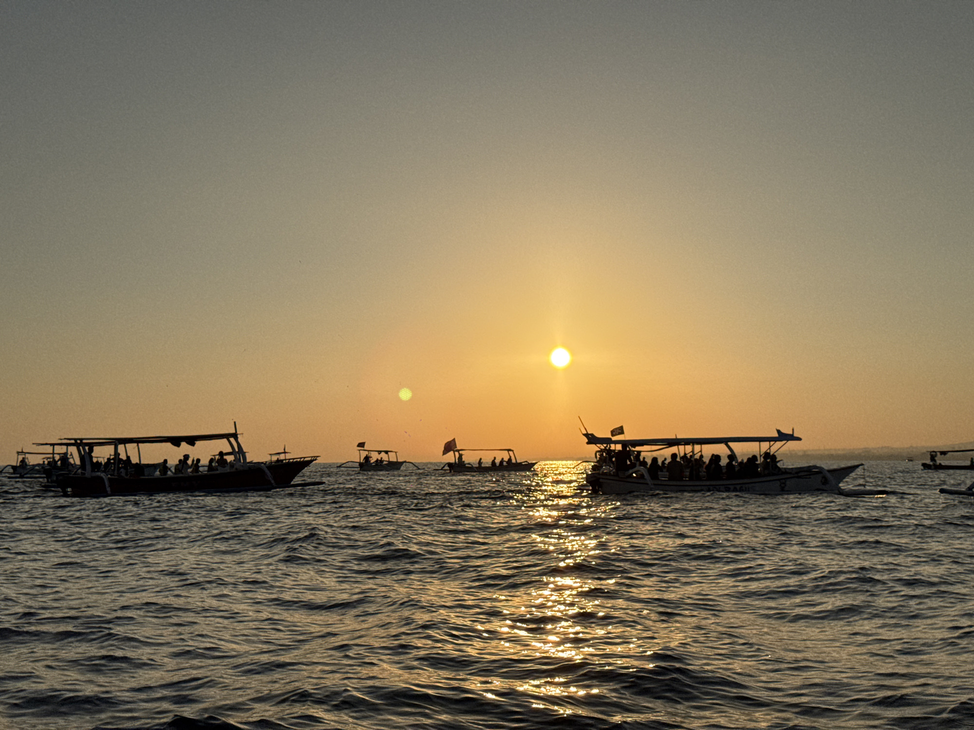 A wide shot captures several boats filled with people on the ocean during sunset. The sun, a bright, circular orb, is positioned slightly right of the frame's center, casting a golden sheen across the rippling water. The sky transitions from a pale yellow near the horizon to a muted gray at the top. Silhouetted against the vibrant sky are multiple traditional outrigger boats, each with a canopy providing shade for its passengers. The boats are spread out across the frame, with the largest and most prominent boat in the foreground, on the left side of the image. This boat is packed with individuals, their forms indistinct in the dim light. Other boats, also carrying groups of people, are visible further in the distance, some with small flags. The overall scene suggests a leisurely outing or a tour group enjoying the sunset at sea, likely in a coastal tourist destination in Indonesia.